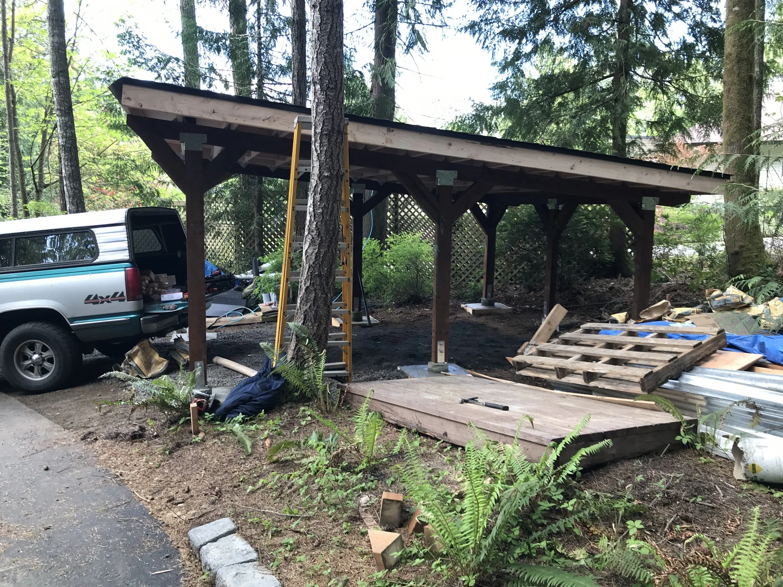 The image shows a construction site in a wooded area where a wooden deck or porch is being built. There is a pickup truck with an open bed filled with construction materials, a yellow ladder, and various tools and materials scattered around. The stru
