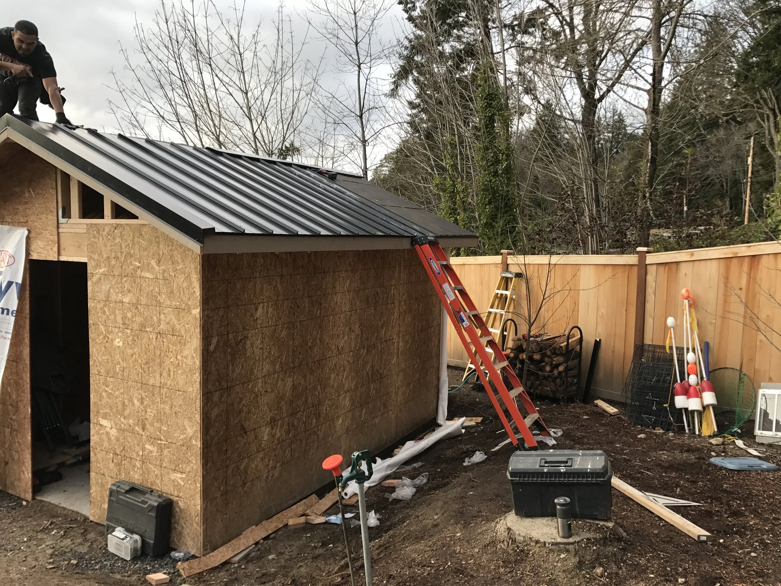 Construction site of a small shed or house, with a worker installing a metal roof, surrounded by tools and wood materials, in an outdoor area with trees.