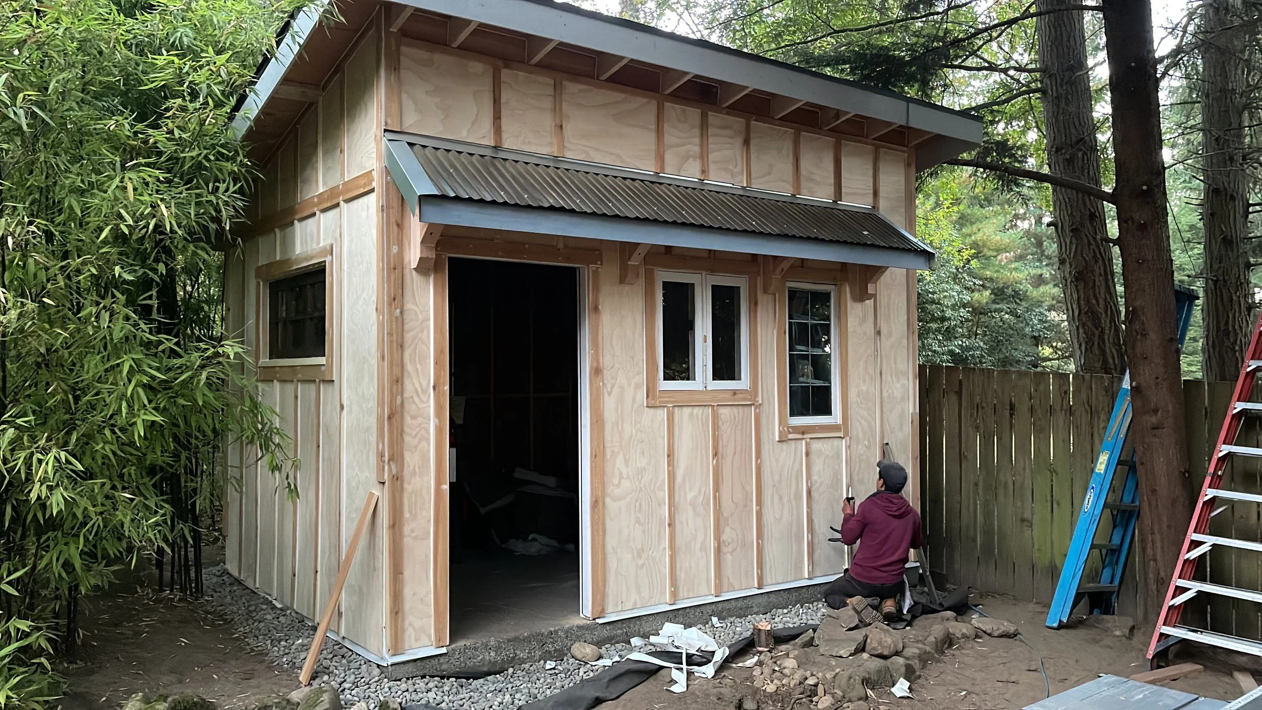 A two-story wooden house under construction surrounded by trees. The house has exposed wood framing, and the front door and windows are installed. A person in a burgundy jacket is kneeling outside, working near the foundation. Tools and materials are