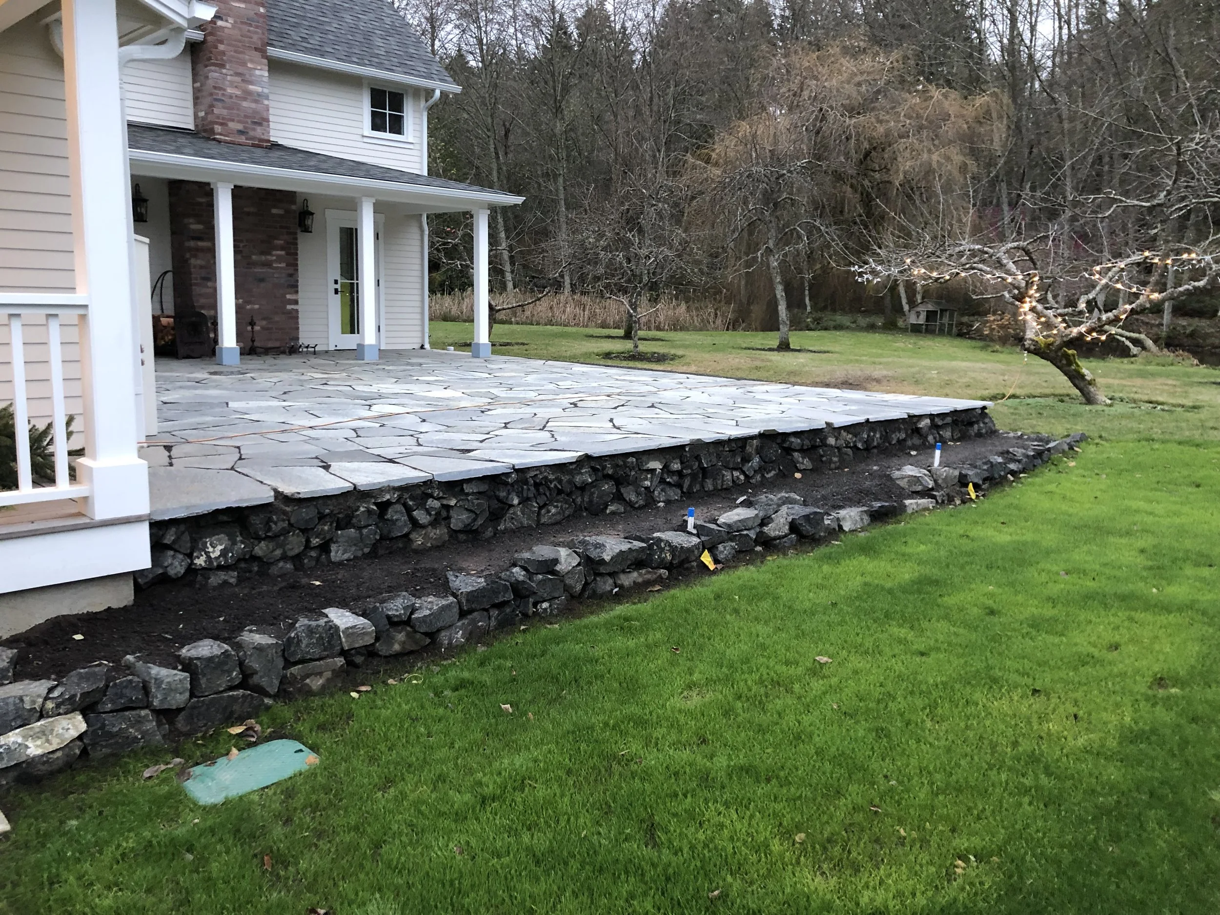 Backyard patio with stone flooring and a stone retaining wall, adjacent to a house with beige siding and white trim, and a backyard with leafless trees and a grassy lawn.