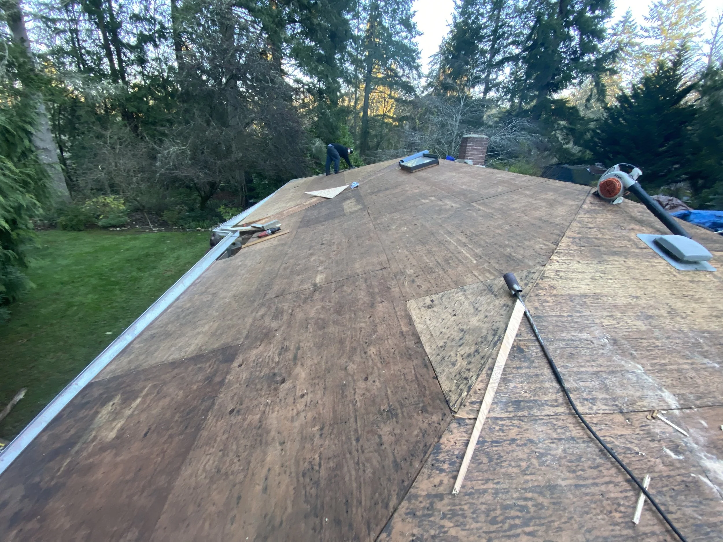 Two workers on a house roof with tools, surrounded by trees, installing or repairing roofing shingles.