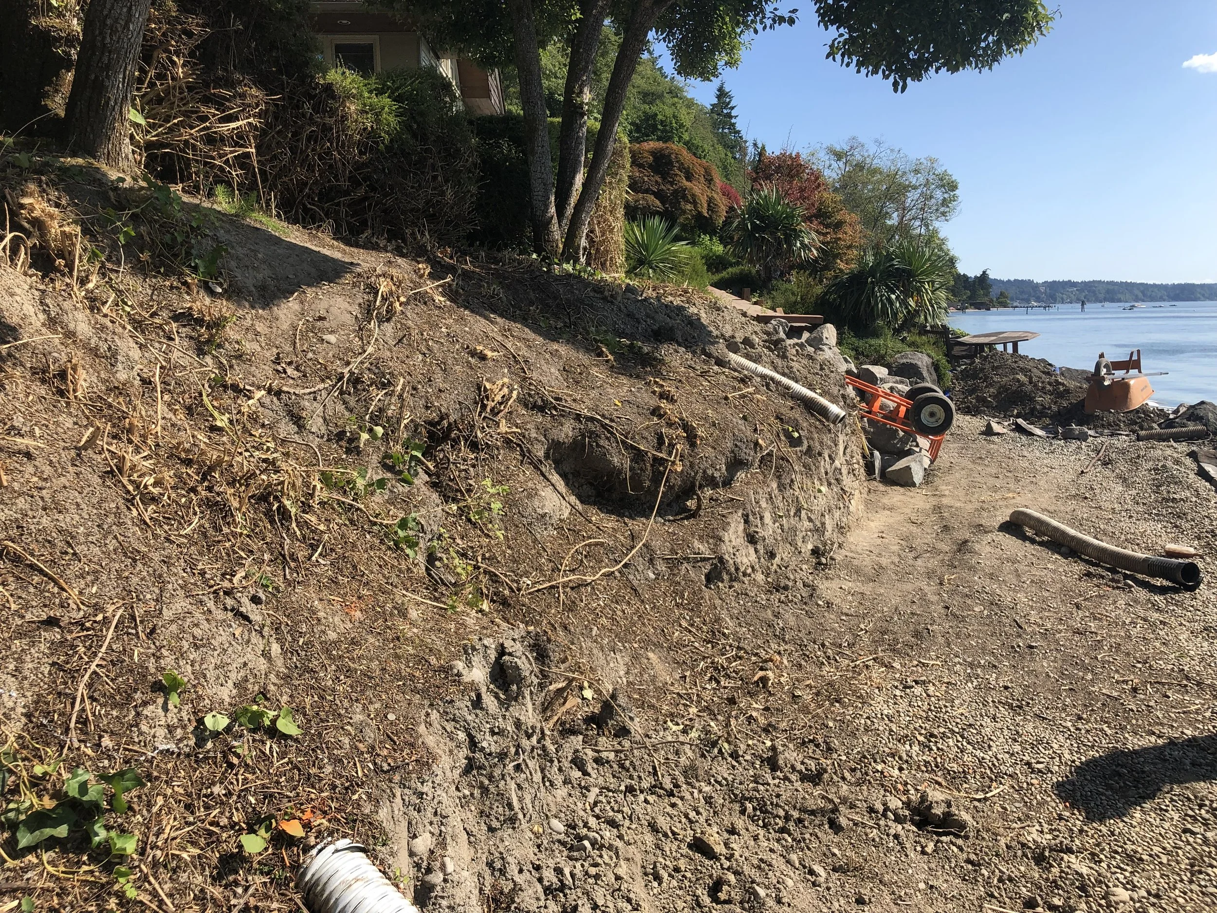 Eroded shoreline with exposed soil, rocks, and construction pipes, next to a body of water with boats and a pier in the background, under a clear blue sky.