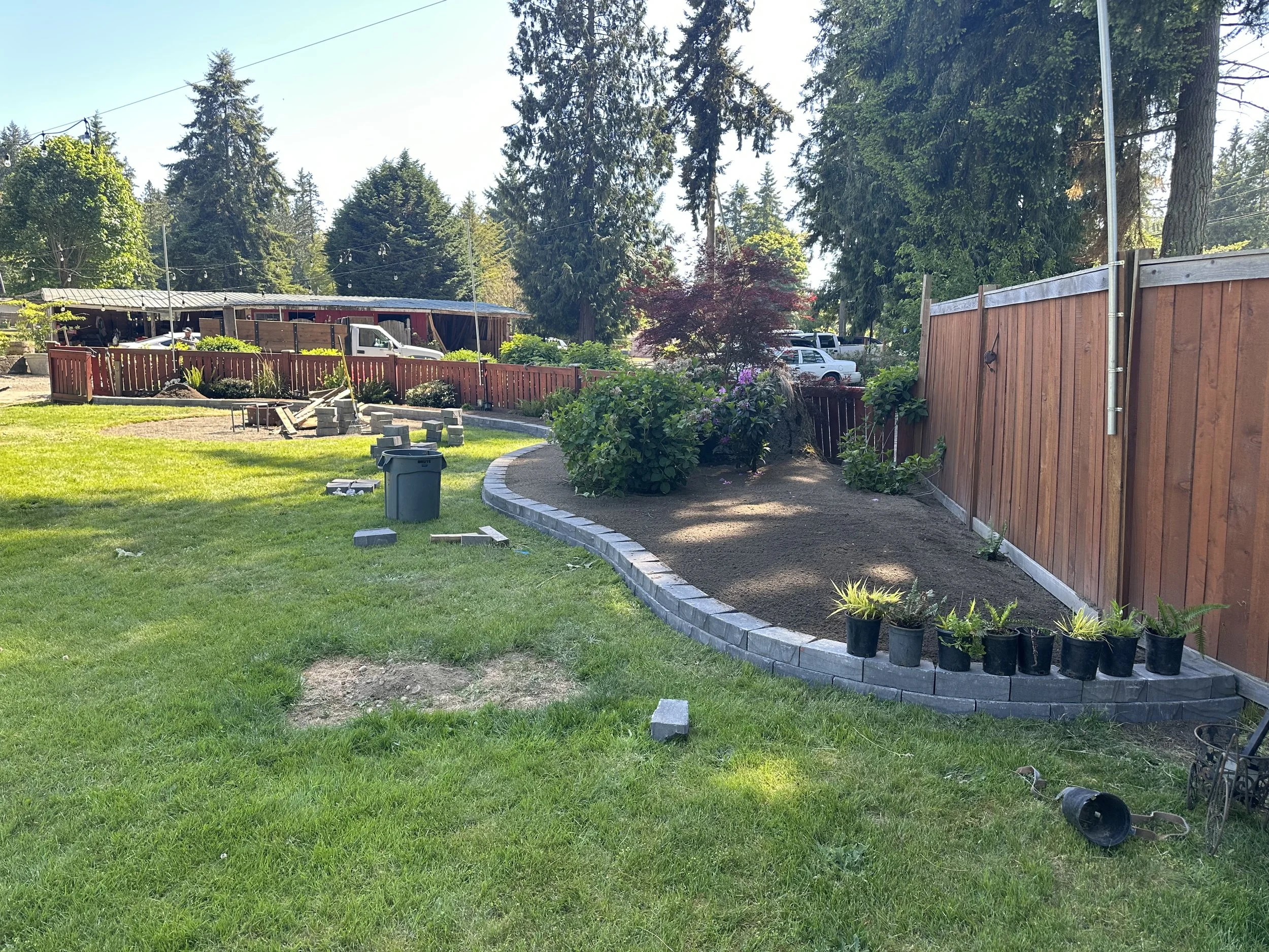 A backyard with a green lawn, a flower bed, a wooden fence, and trees in the background. There are construction blocks and potted plants near the fence, and cars are parked behind it. The sky is clear and sunny.