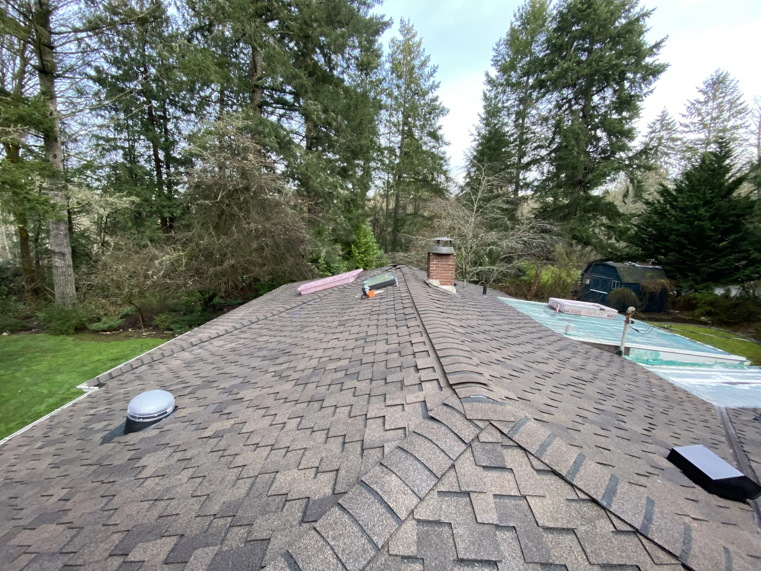 View of a residential rooftop with asphalt shingles, surrounding trees, and some construction materials on the roof, including a pink insulation board and a black container, under a cloudy sky.