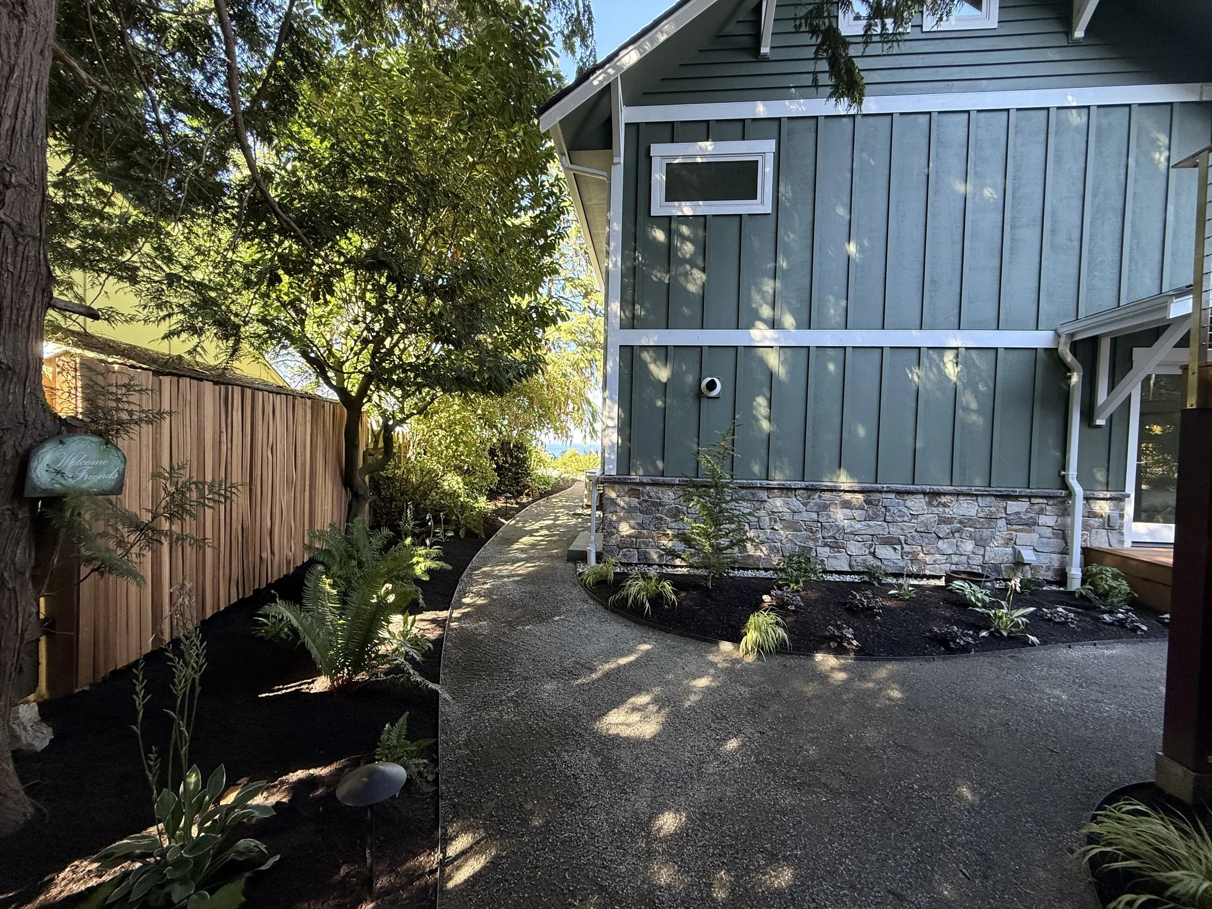 A backyard garden with a wooden fence, green shrubbery, and trees, next to a house with blue siding, a small window, and a stone foundation, with a paved walkway and garden lighting.