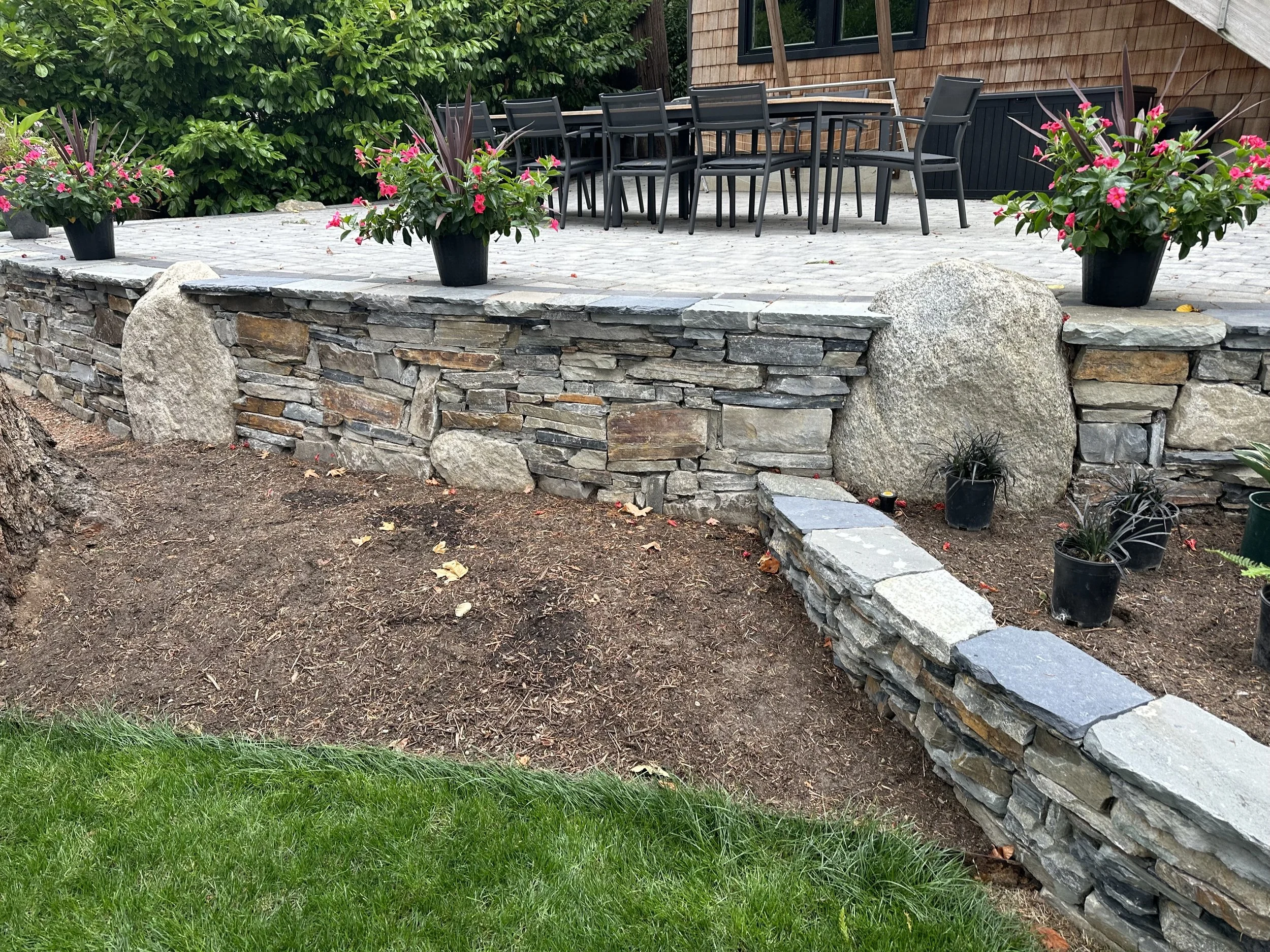 A backyard patio with a stone wall, potted pink flowers, a stack of black patio chairs, and a wooden house in the background.