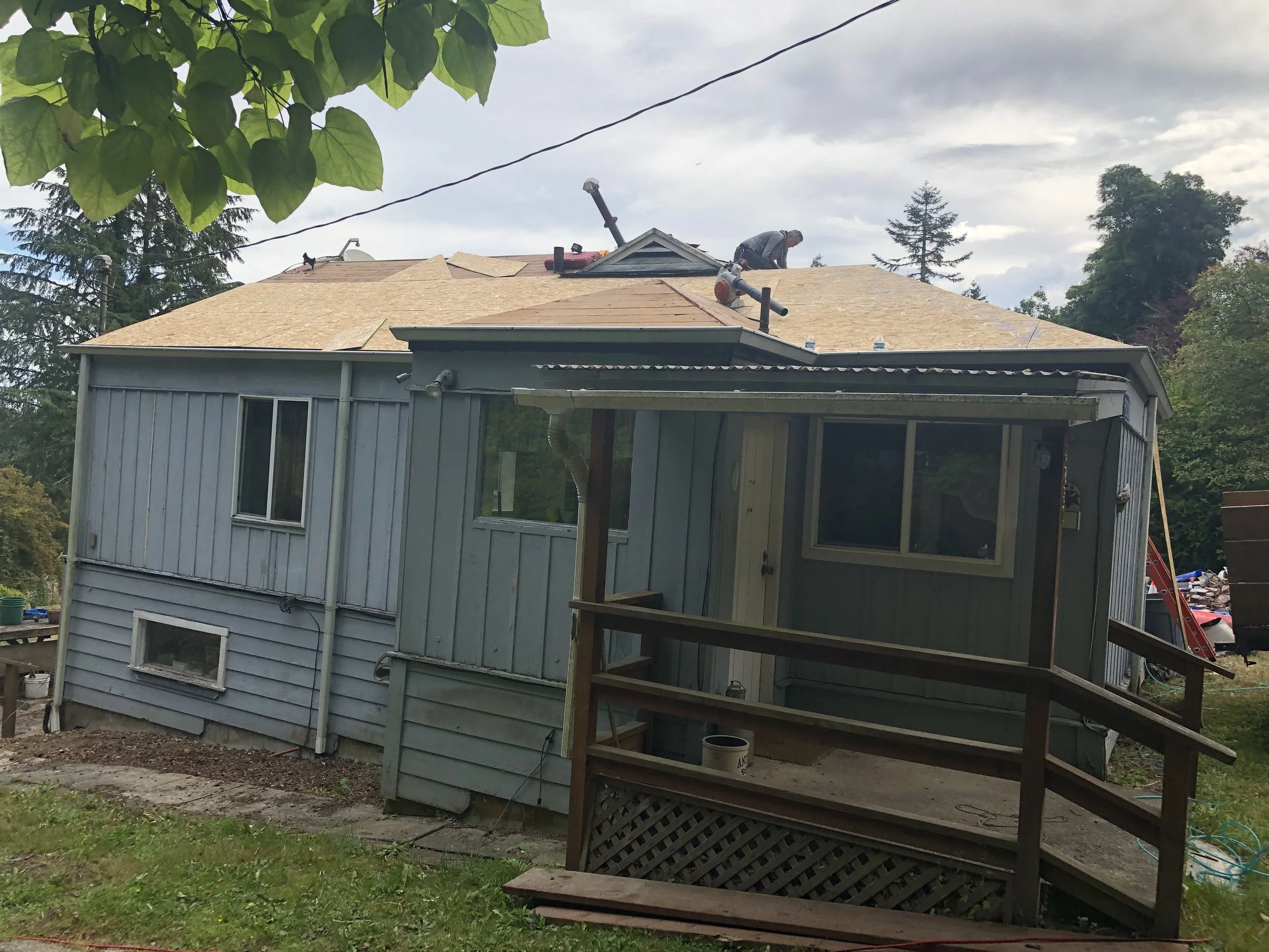 A house under renovation with workers on the roof, which is partially covered with plywood sheets. The house has light-colored exterior siding and a screened porch. There are trees and a cloudy sky in the background.