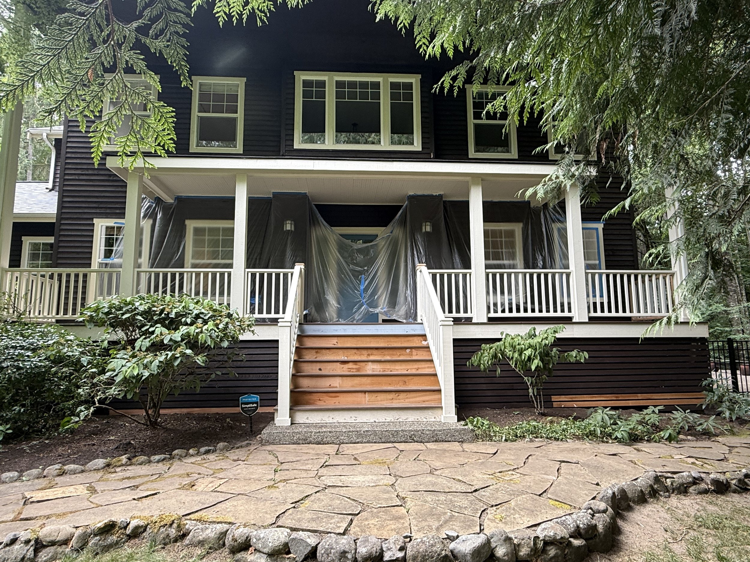 Front view of a two-story house under construction, with black siding, white trim, and multiple windows; a staircase leads to the porch, which is covered with black plastic; surrounding greenery includes bushes, trees, and a brick patio.