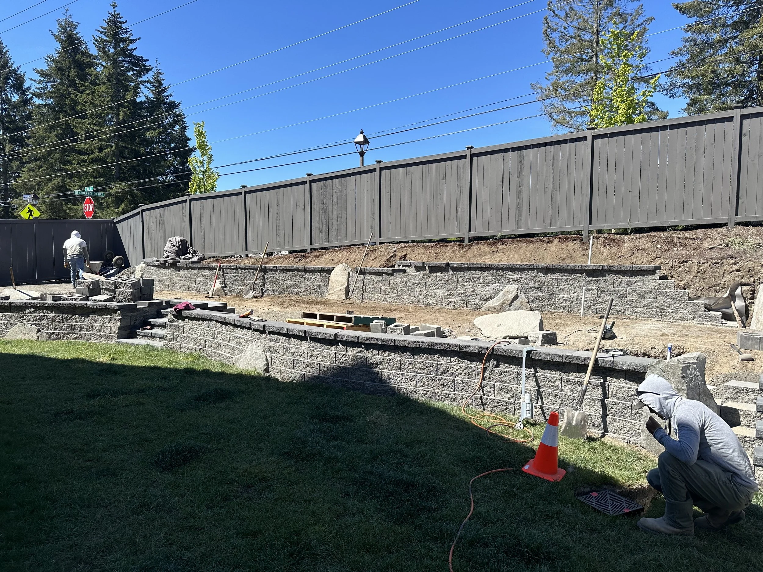 Construction workers installing a wooden fence on a hillside yard with trees and power lines in the background.