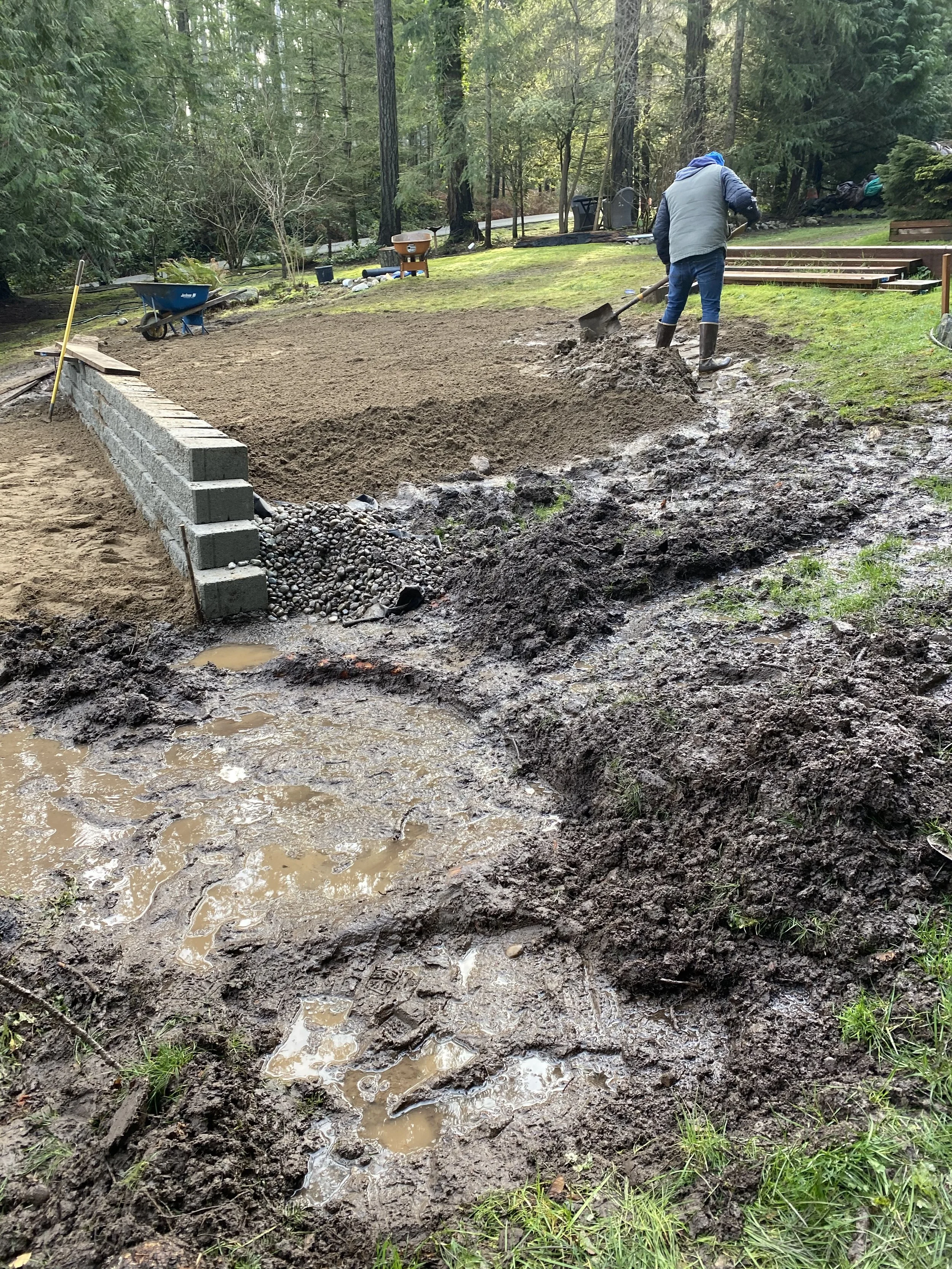 Person shoveling wet soil on a muddy area surrounded by trees and grass, with construction materials and tools nearby.