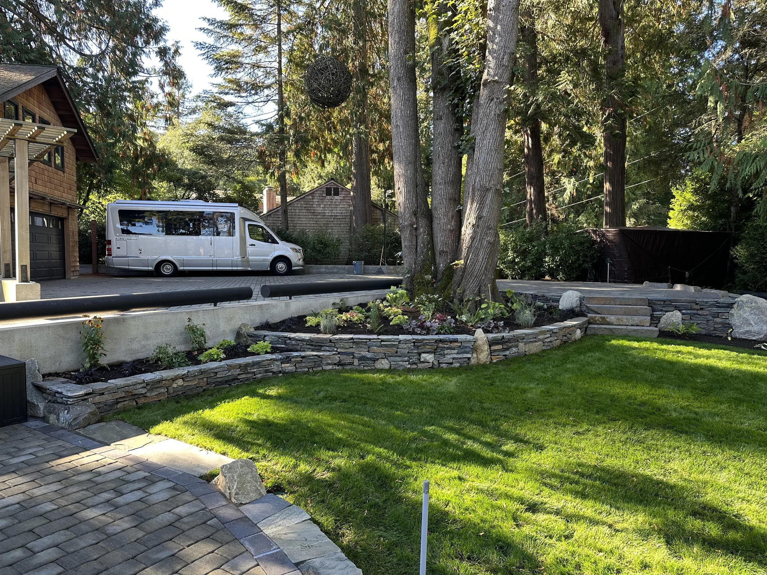 Residential backyard with a paved patio, a small grassy yard with flower beds, a large tree, a white van parked, and a house with wooden siding in the background.