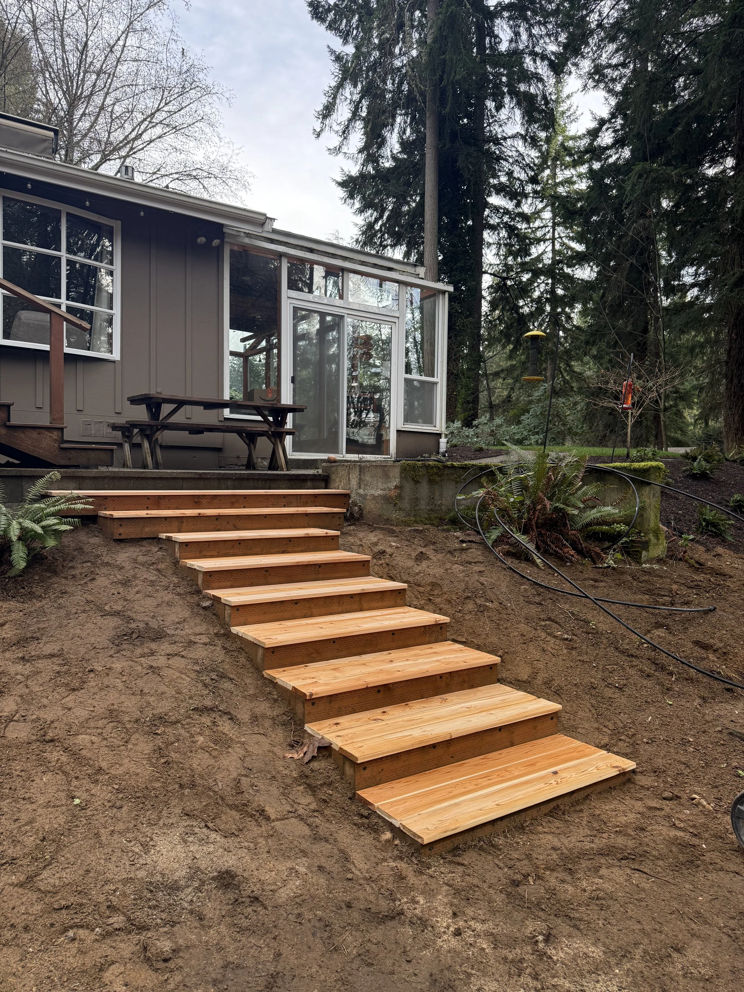 Wooden steps leading up to a house with a screened porch, surrounded by trees and outdoor plants.