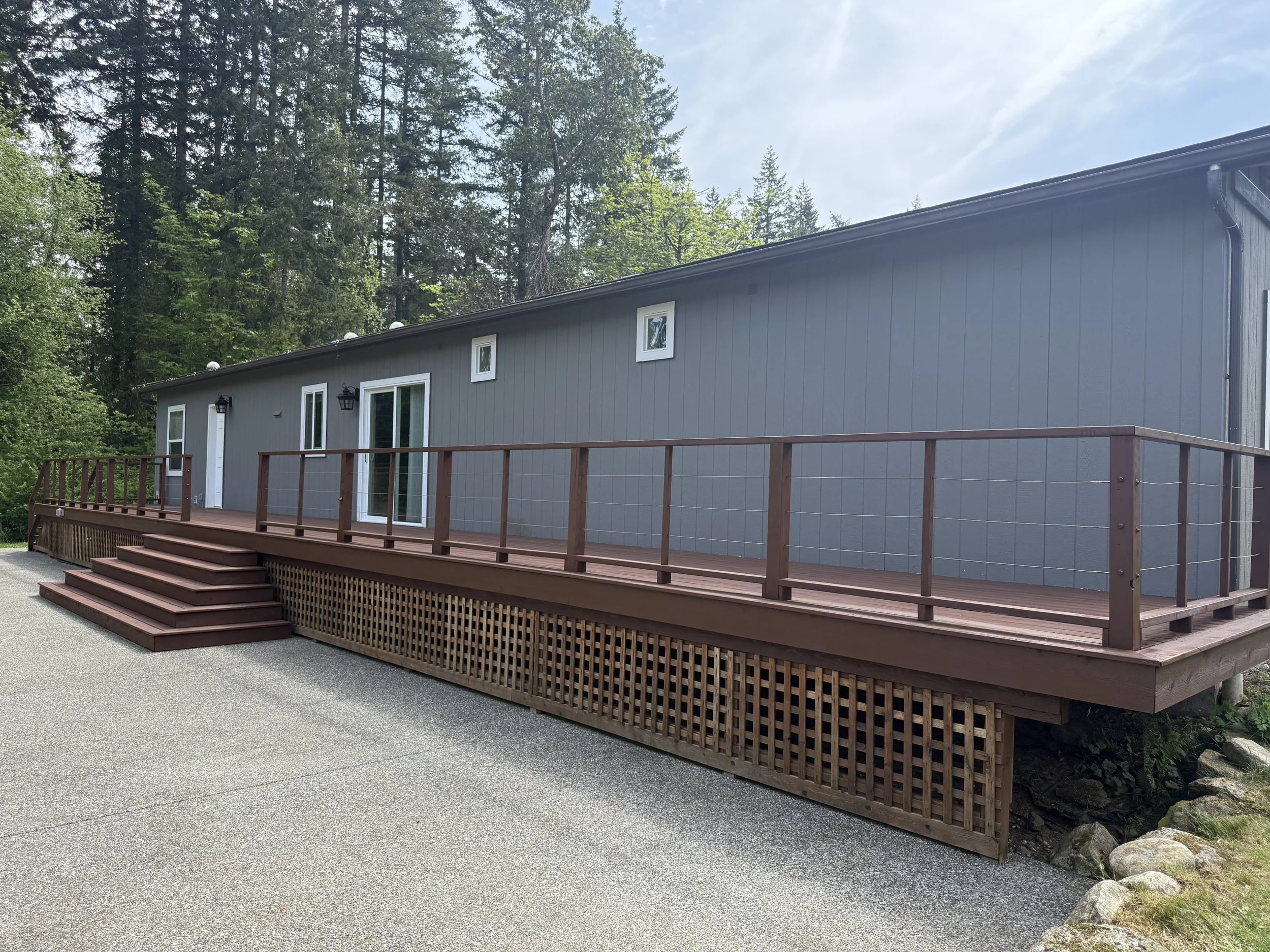 Gray house with white trim and several small windows, a wooden front deck with stairs, black exterior wall lights, and a wooded background with tall trees and a blue sky.
