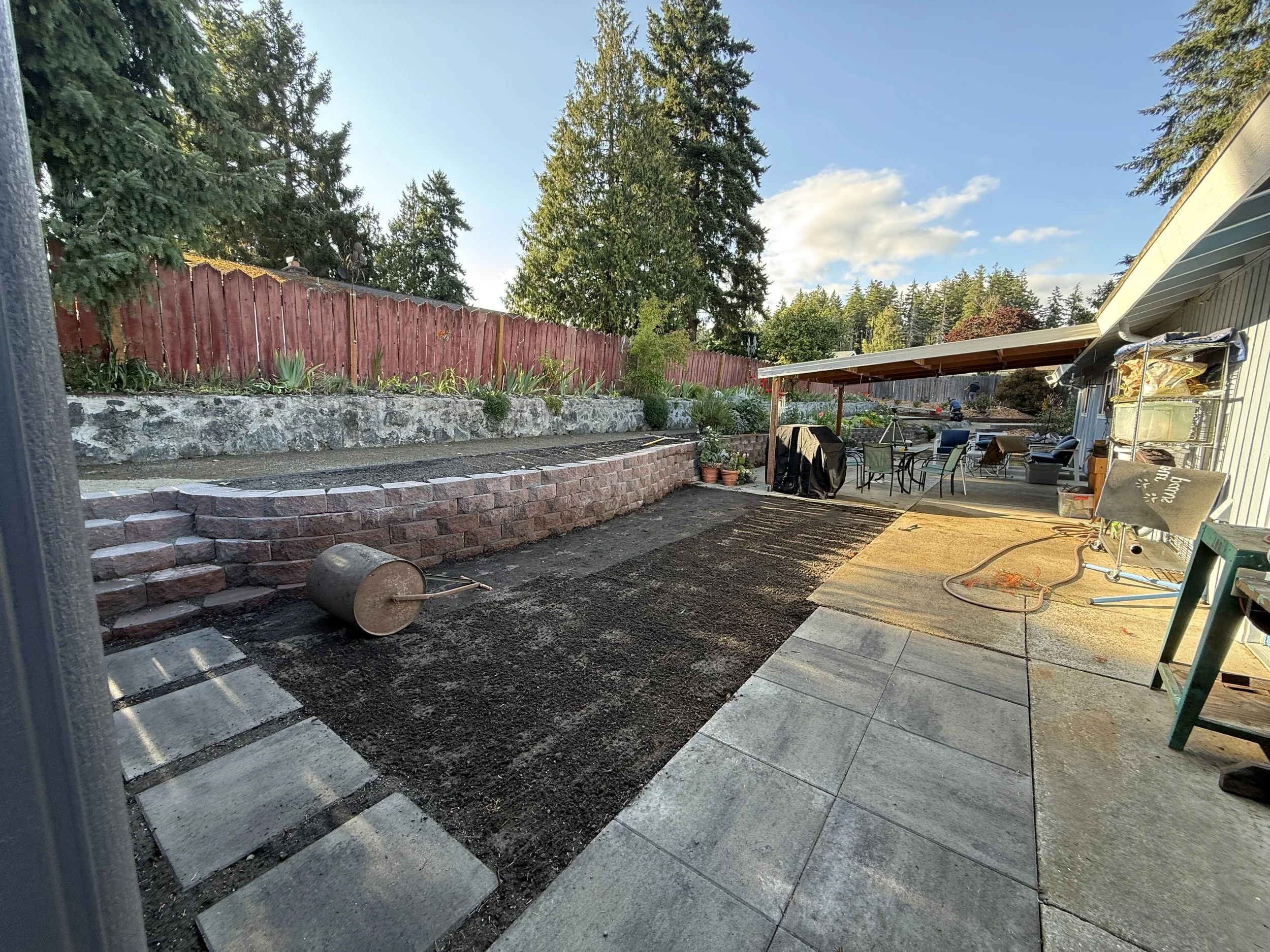 A backyard patio with a covered seating area, potted plants, a red wooden fence, and large evergreen trees in the background.