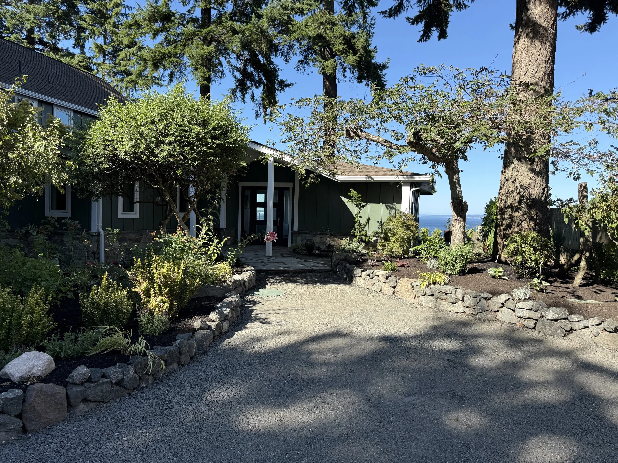A house with dark green walls and a gray roof surrounded by trees and a garden with rocks and plants, overlooking a body of water in the distance.