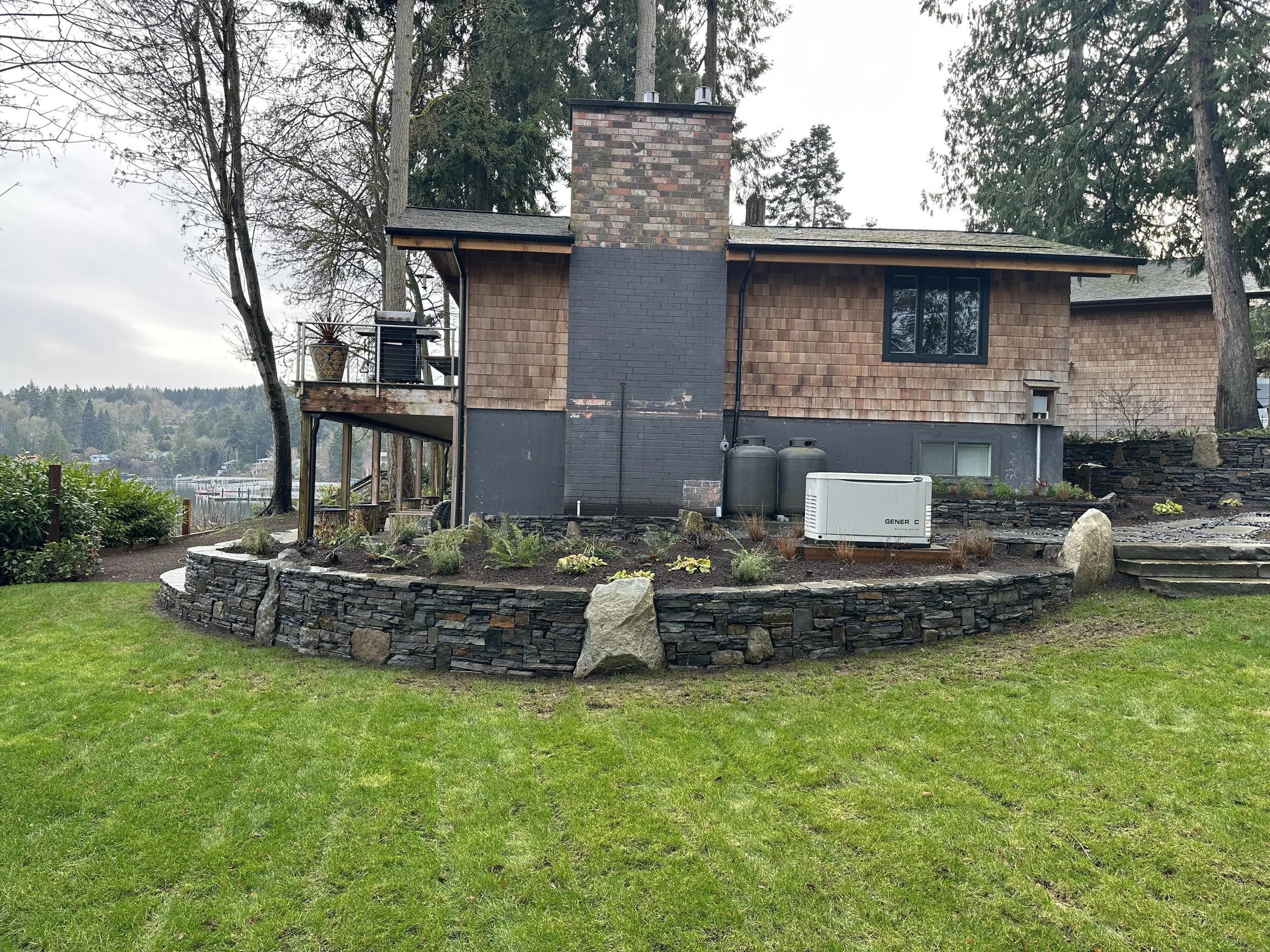 A house with a brick chimney, brown shingle siding, and black window frames, surrounded by tall trees and a landscaped yard with a stone border and green lawn.