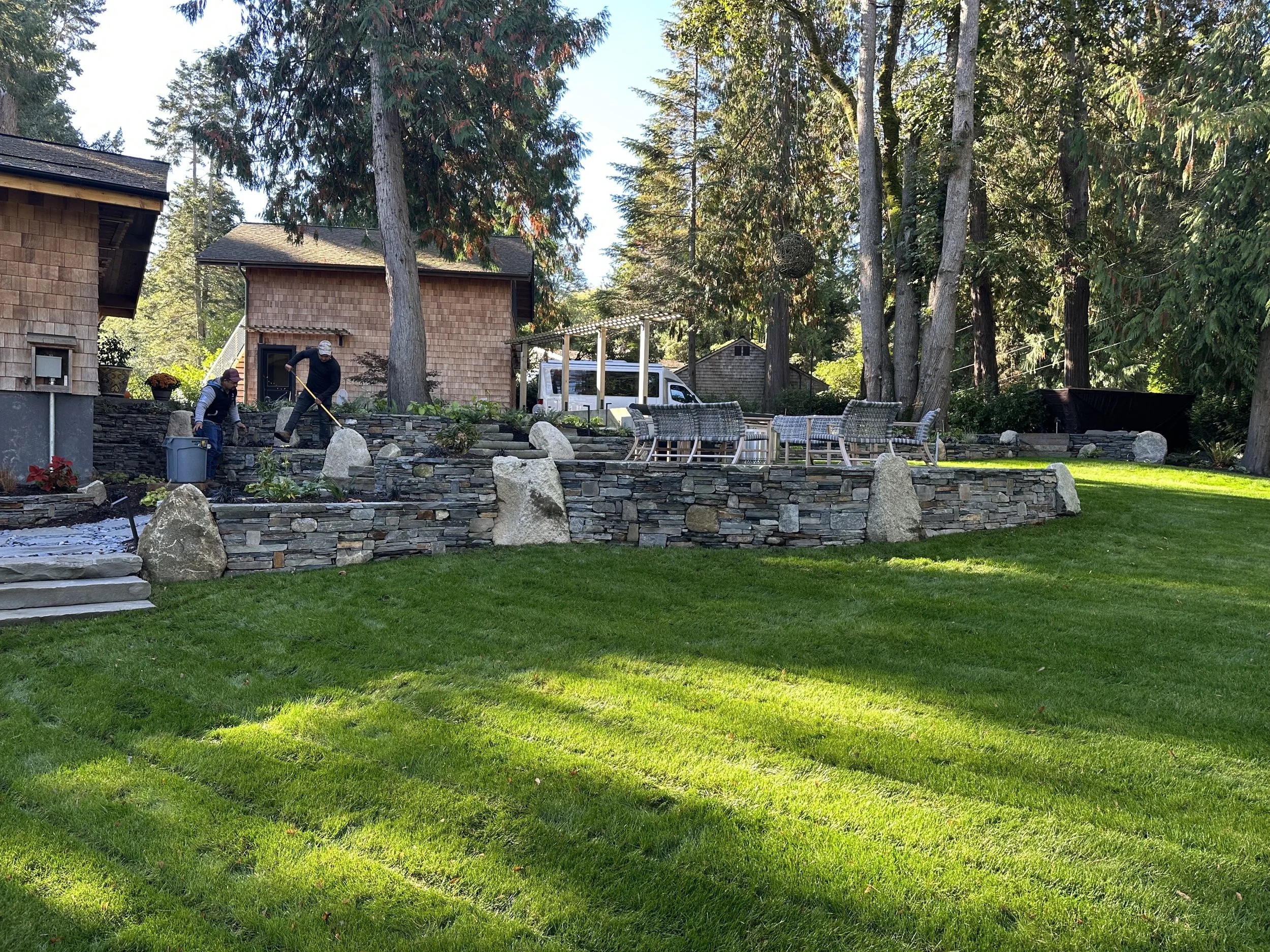 Two men working on a stone garden wall in a backyard, surrounded by tall trees and sunshine, with patio chairs and a white van nearby.