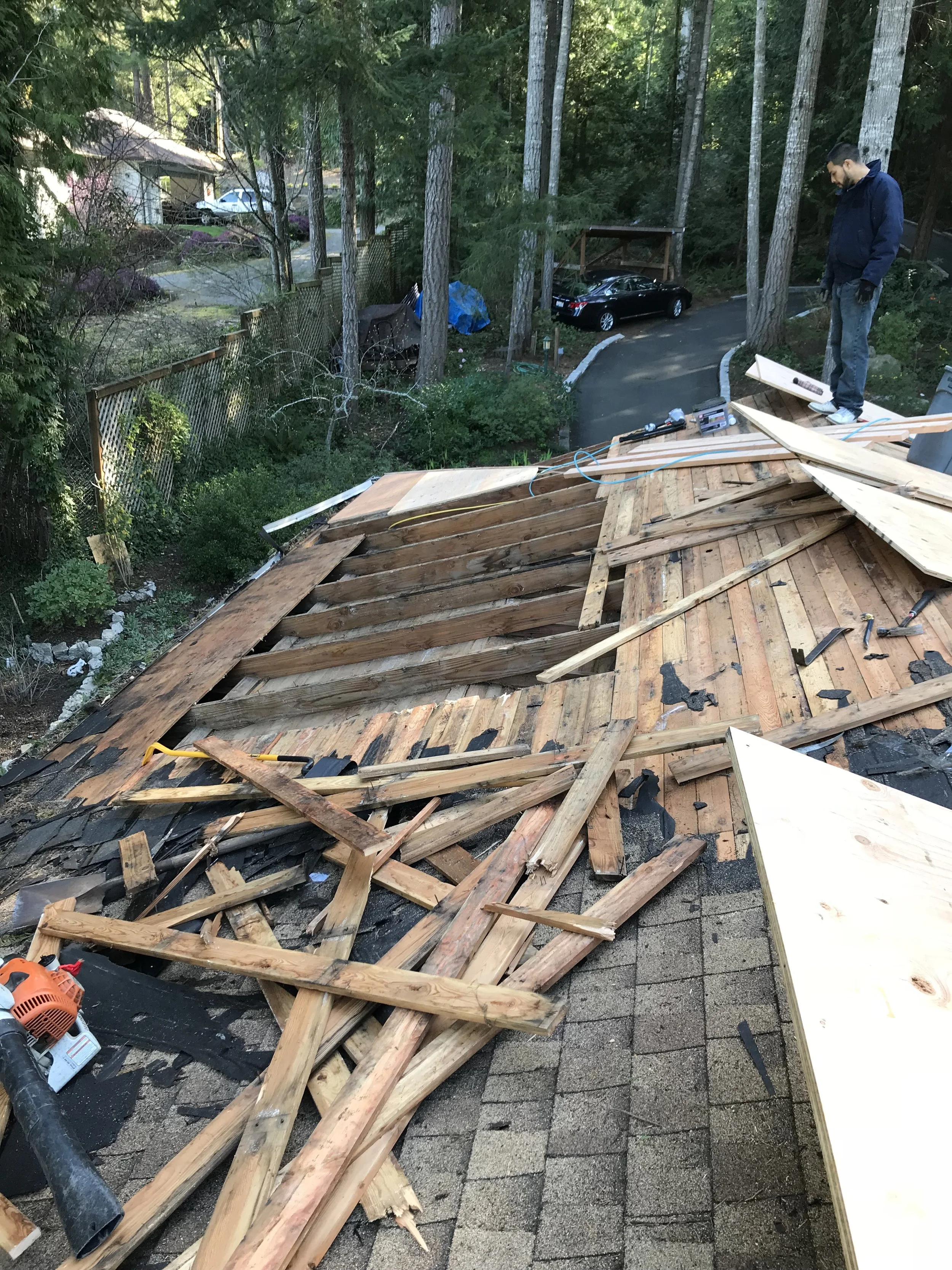 Rear view of house with partially constructed roof; worker standing on roof framing, surrounded by construction tools and materials, with trees and parked cars in the background.