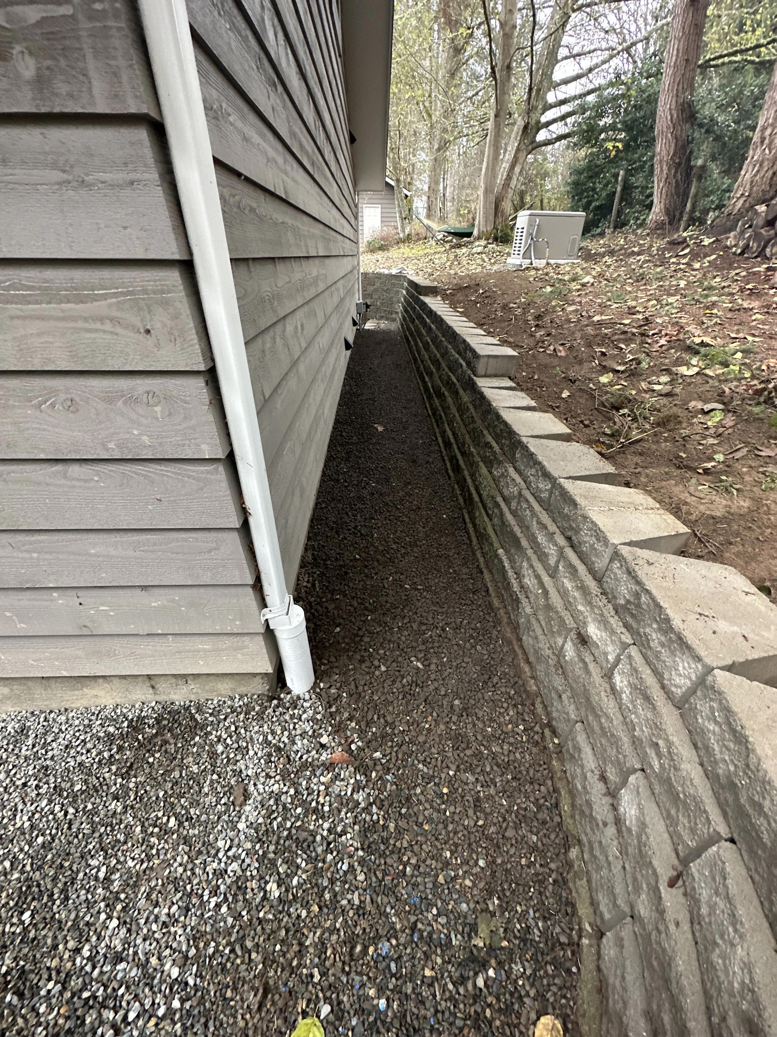 Side of a house with vinyl siding, showing a gravel walkway with a separate gravel bed, a brick retaining wall, and trees in the yard.