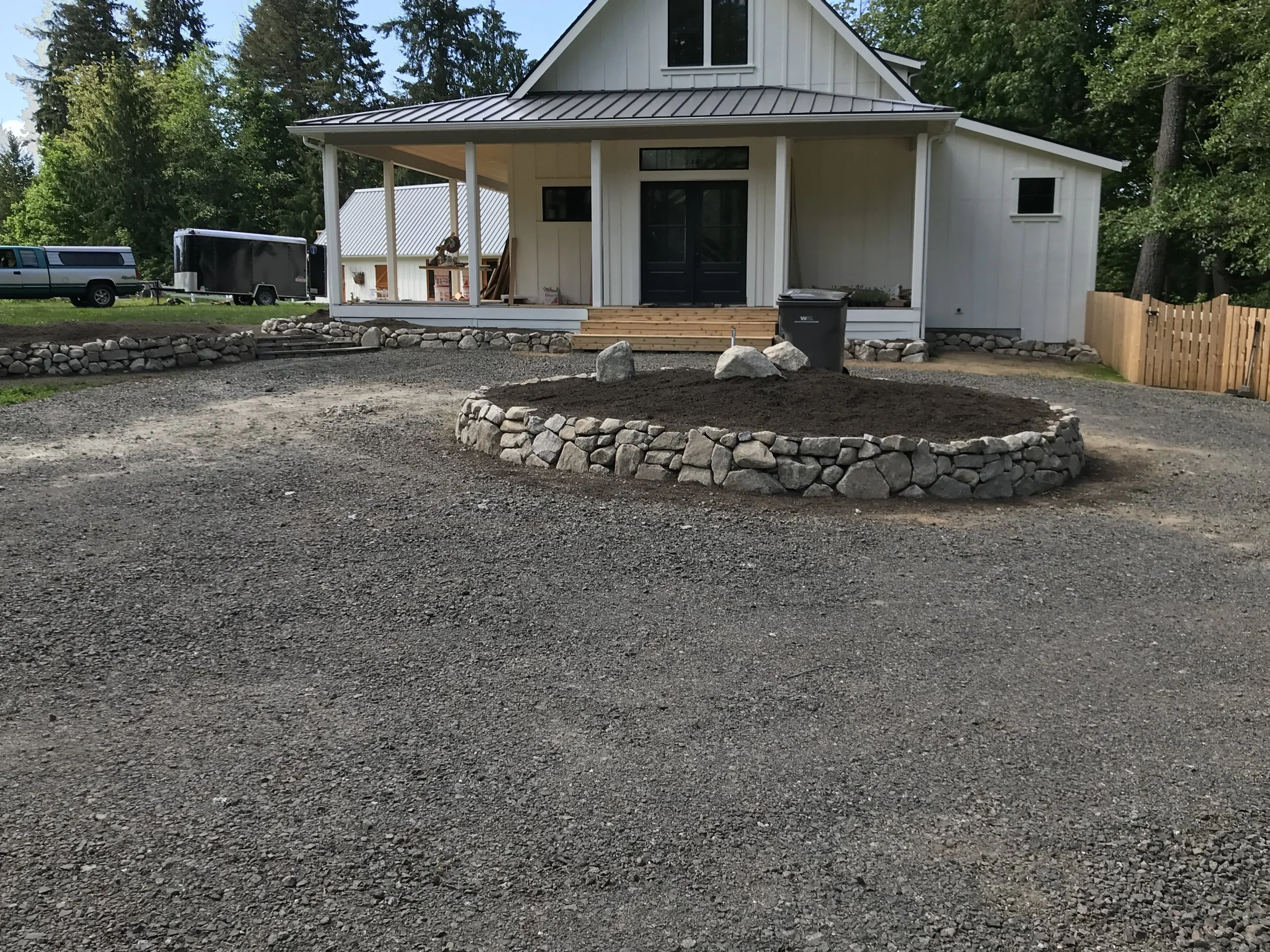 Front view of a white house under construction, with a porch, steps, and a gravel yard, surrounded by trees.