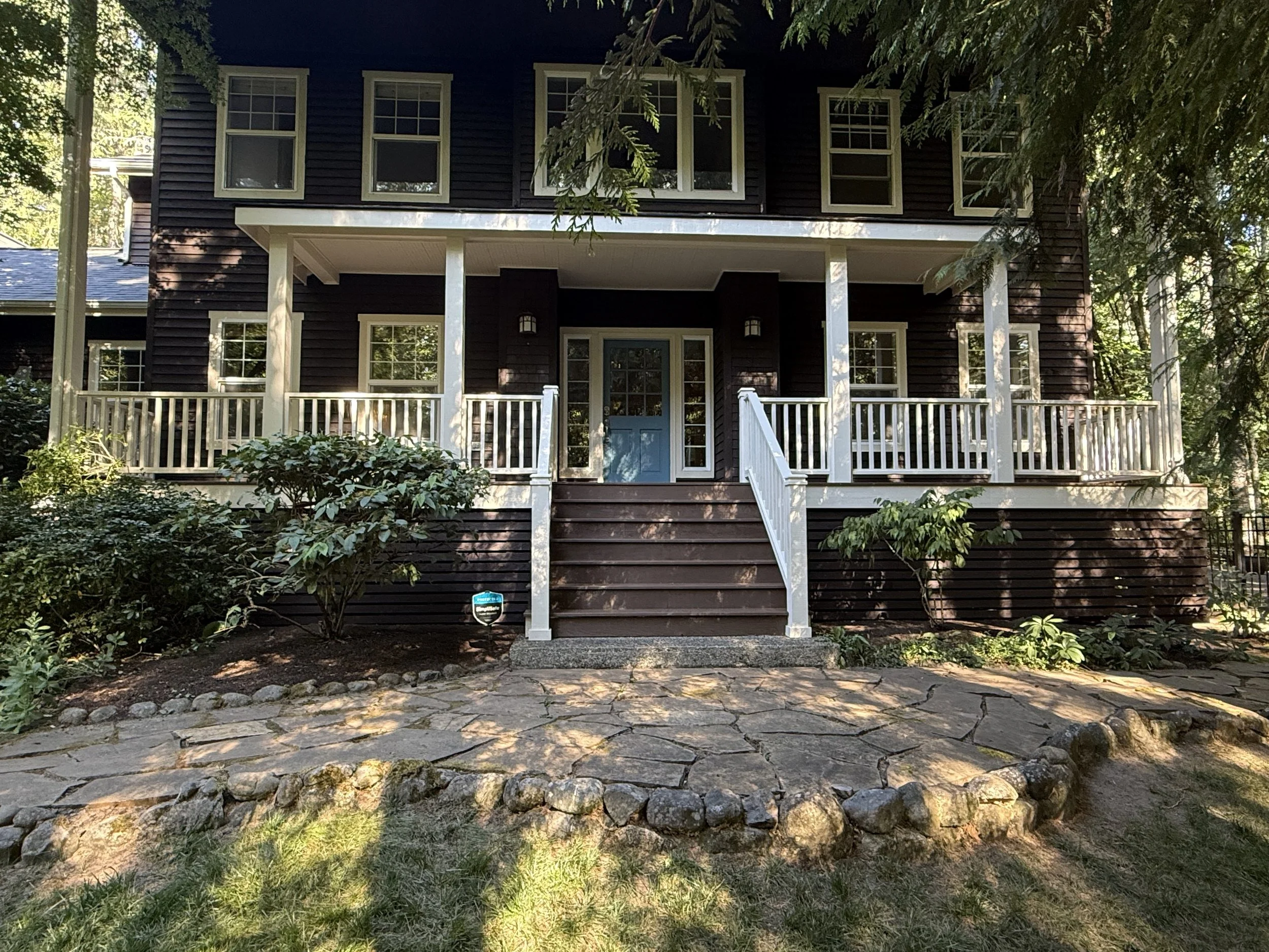Front view of a two-story house with dark brown siding, white window frames, and a white porch railing. The house is surrounded by trees and a stone pathway in the front yard.