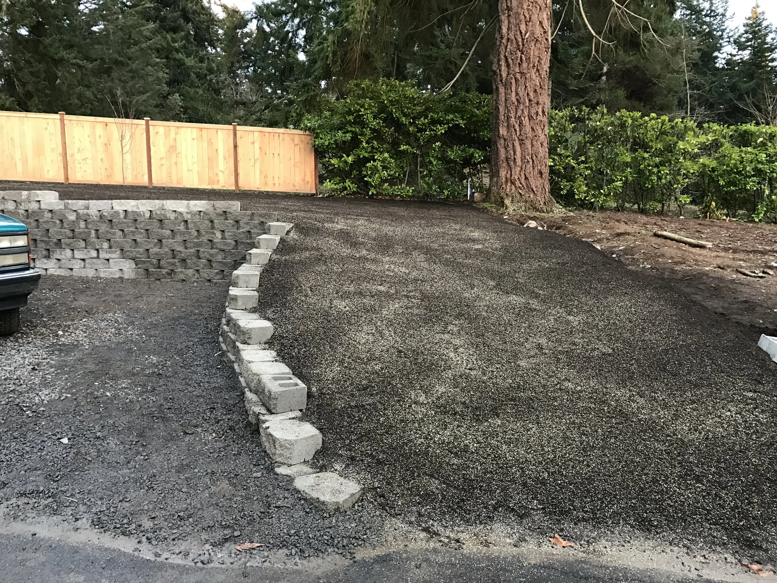A gravel driveway with a short retaining wall of concrete blocks on the left, a large tree on the right, and a wooden fence in the background surrounded by green trees and bushes.