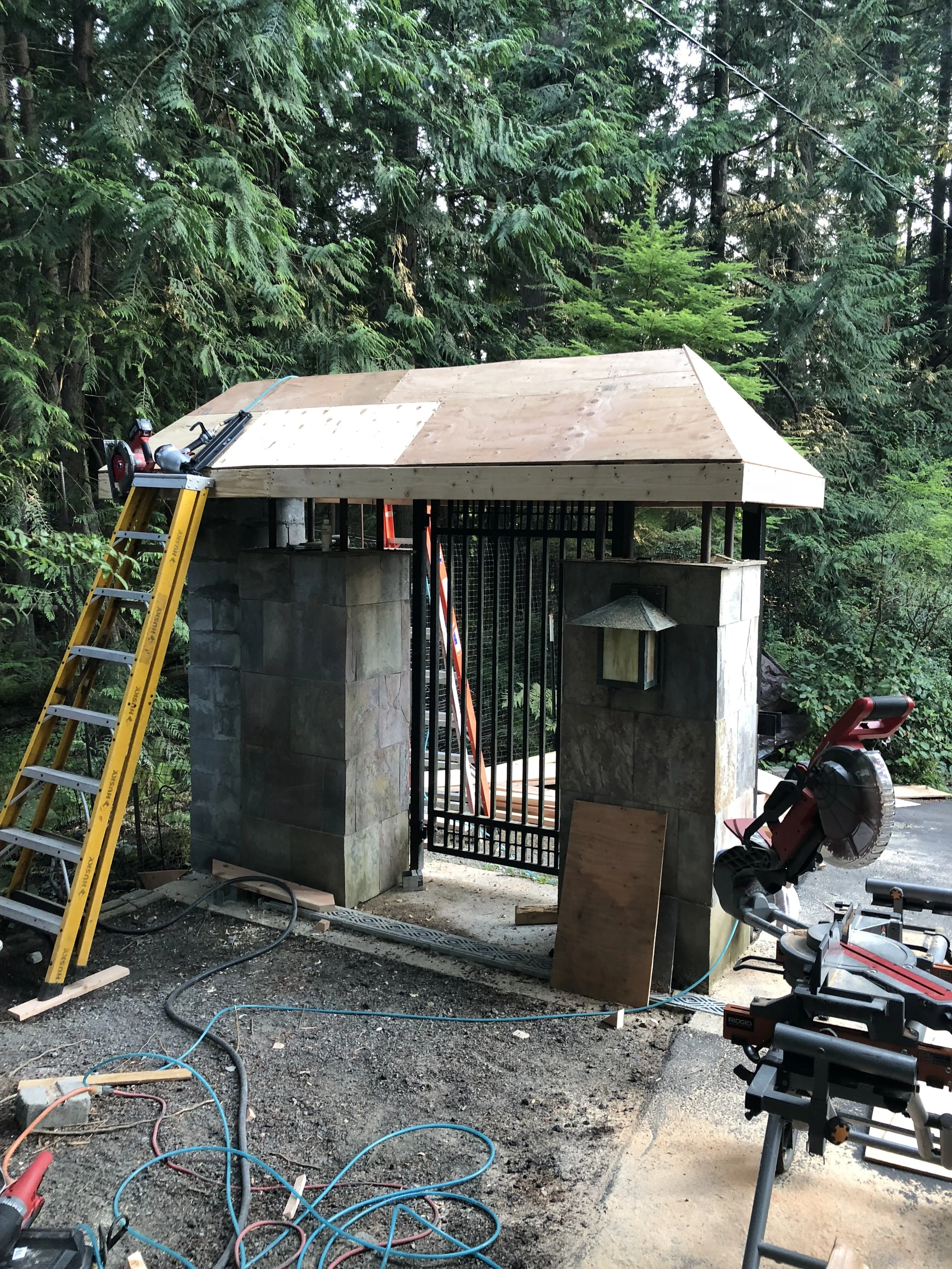 A small structure under construction with a wooden roof, concrete block walls, and a metal gate, surrounded by trees and construction tools including a yellow ladder, power saw, and various cables.