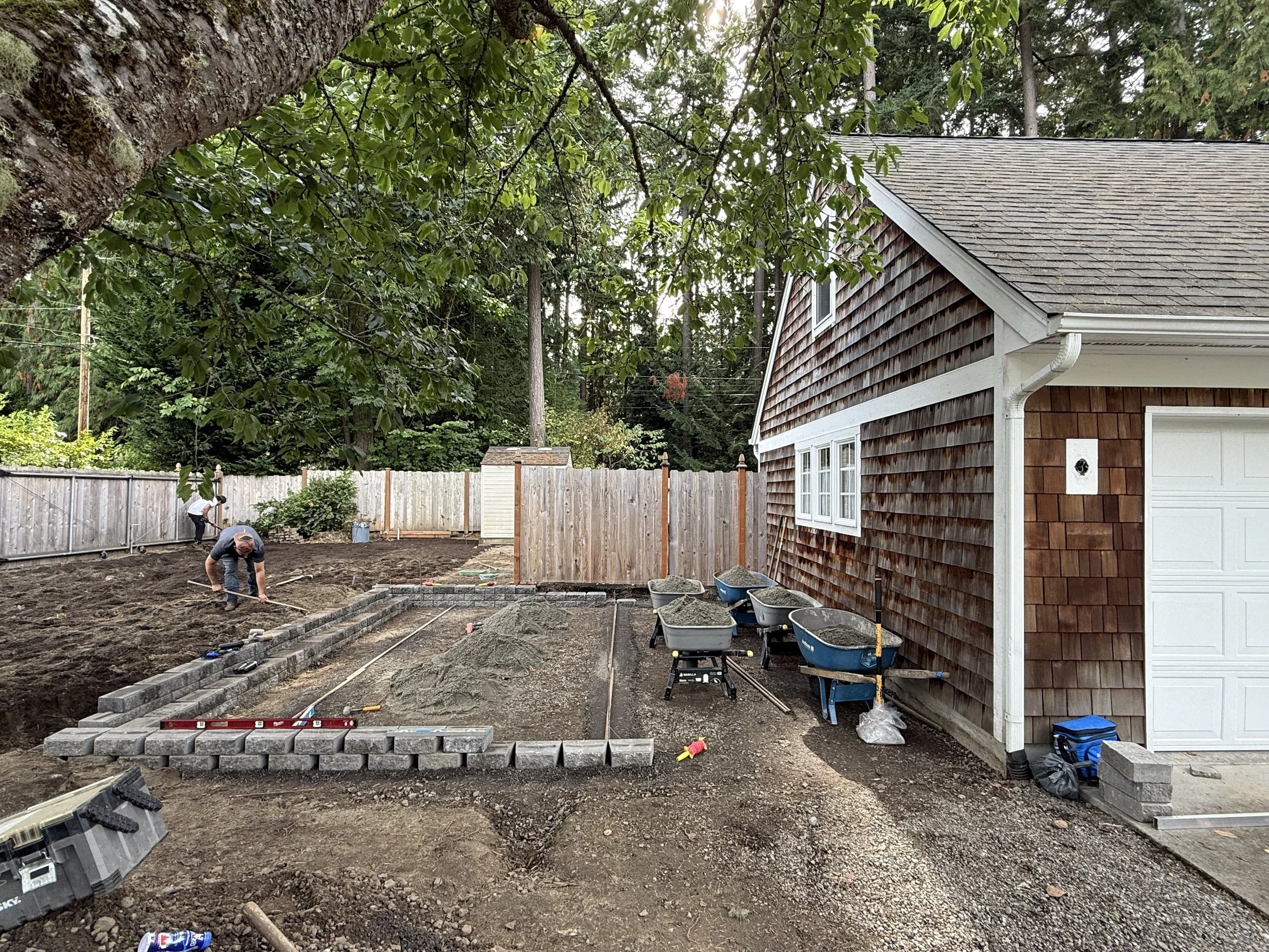 People laying bricks on a construction site in a backyard with a house, trees, and a wooden fence.