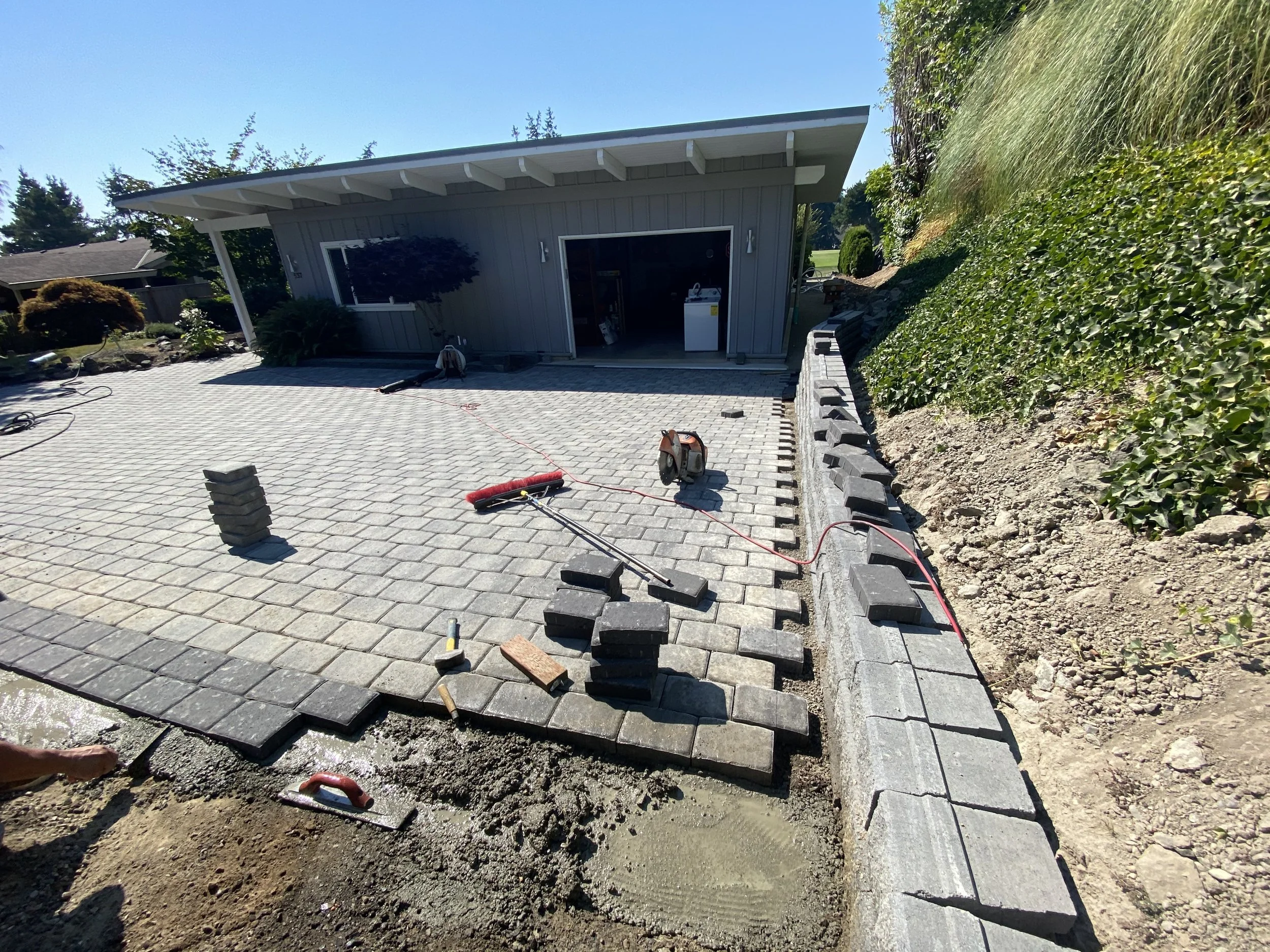 Construction work on a patio with paving stones, tools, and an incomplete border. A small gray building or shed is in the background, and greenery surrounds the area.