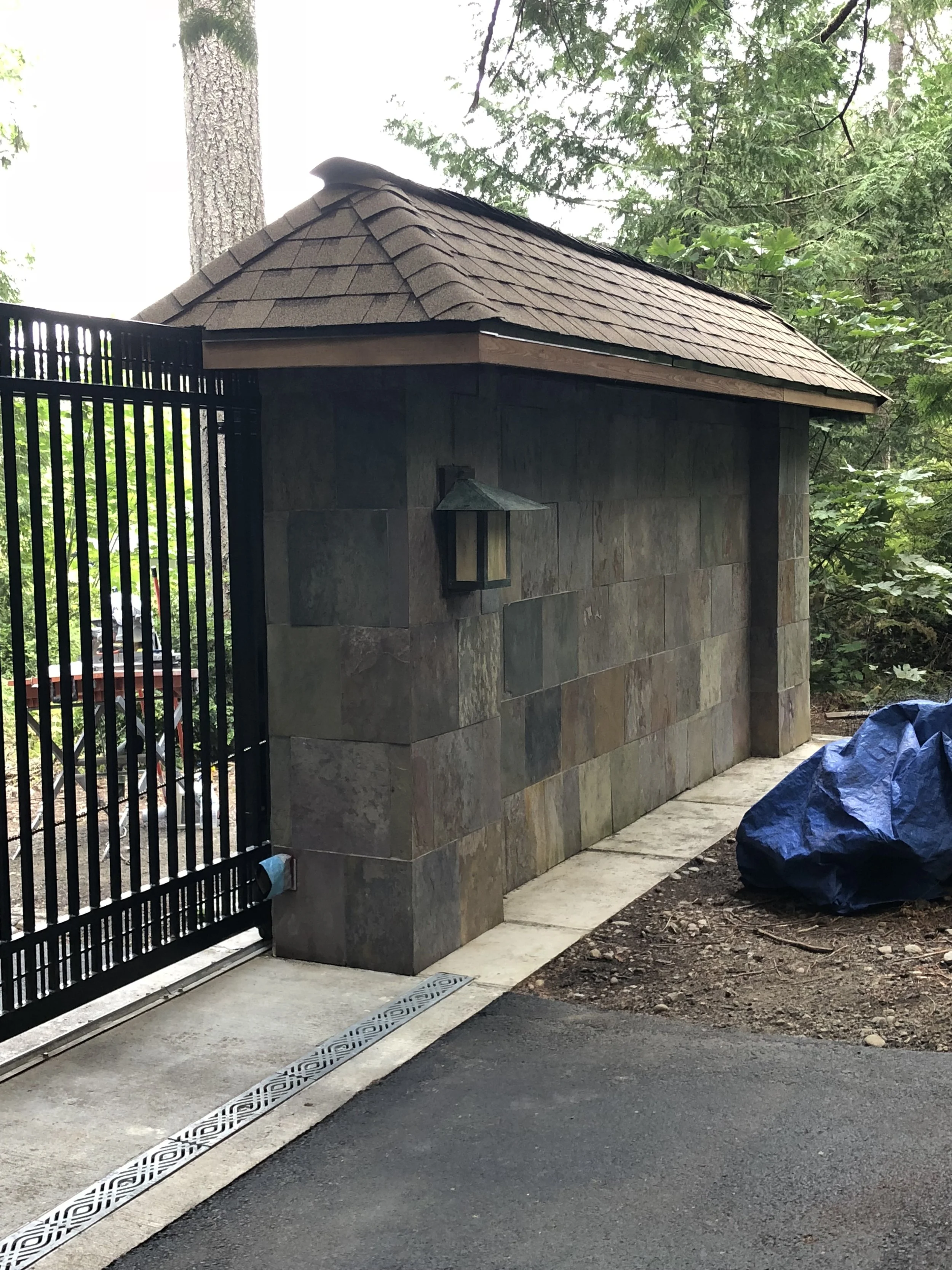 A small structure with a shingled roof and stone wall, located behind a black metal gate, next to a paved pathway with a decorative drain. A blue tarp covers some objects on the ground nearby, with trees and greenery in the background.