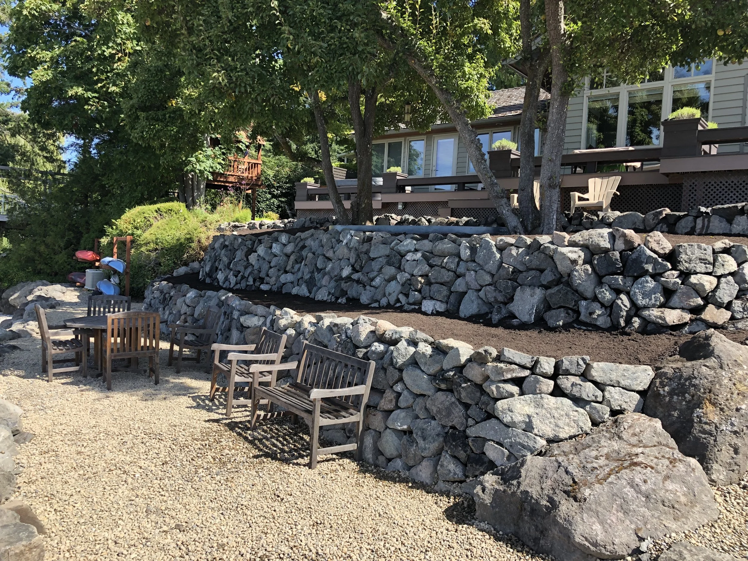 A backyard scene with a stone retaining wall, wooden patio furniture, and a house with large windows and a deck. There are trees providing shade and some buckets and kayaks on the ground.