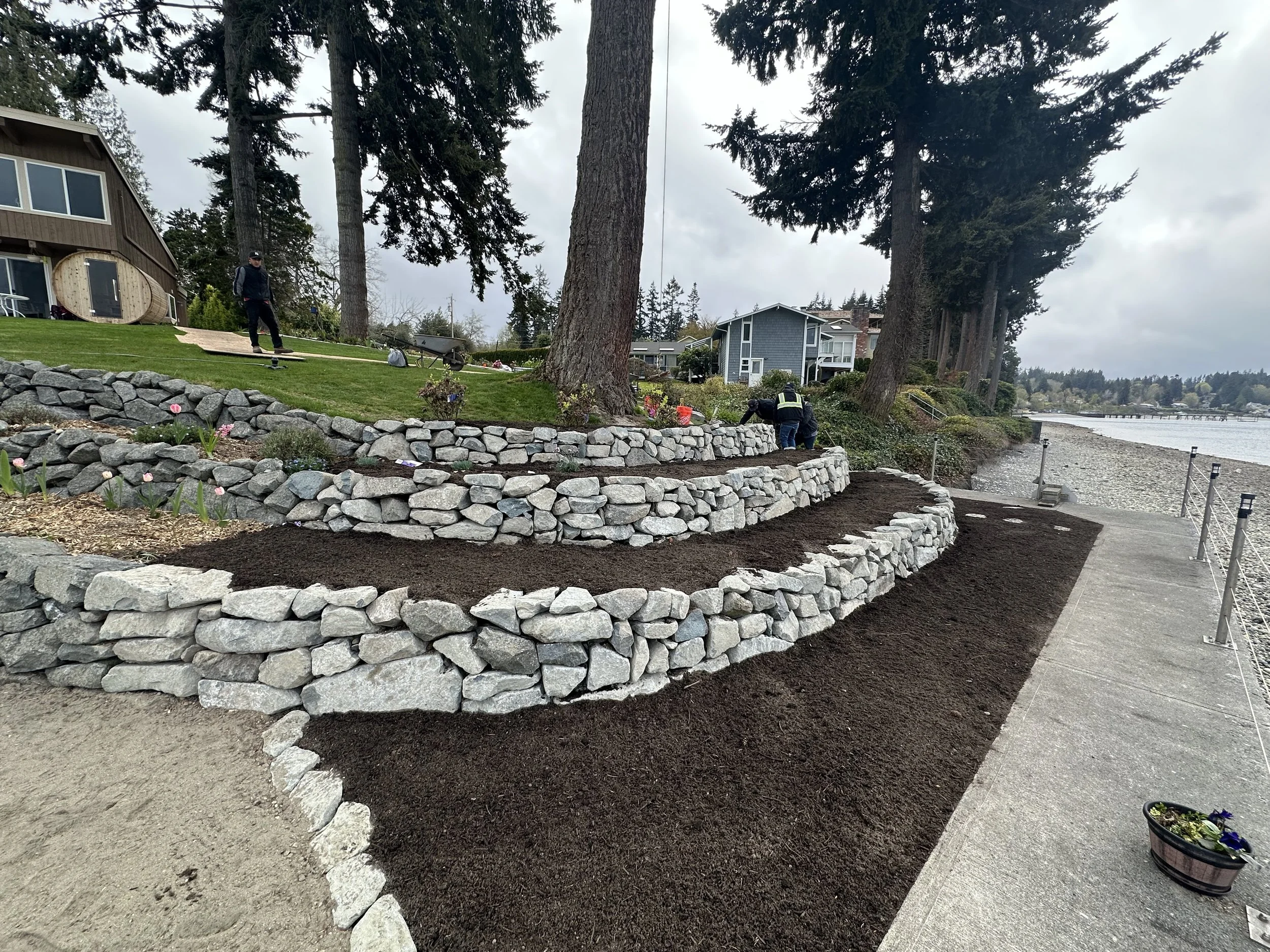 People working on landscaping a backyard garden by a body of water, with stone retaining walls, trees, and residential buildings in the background.
