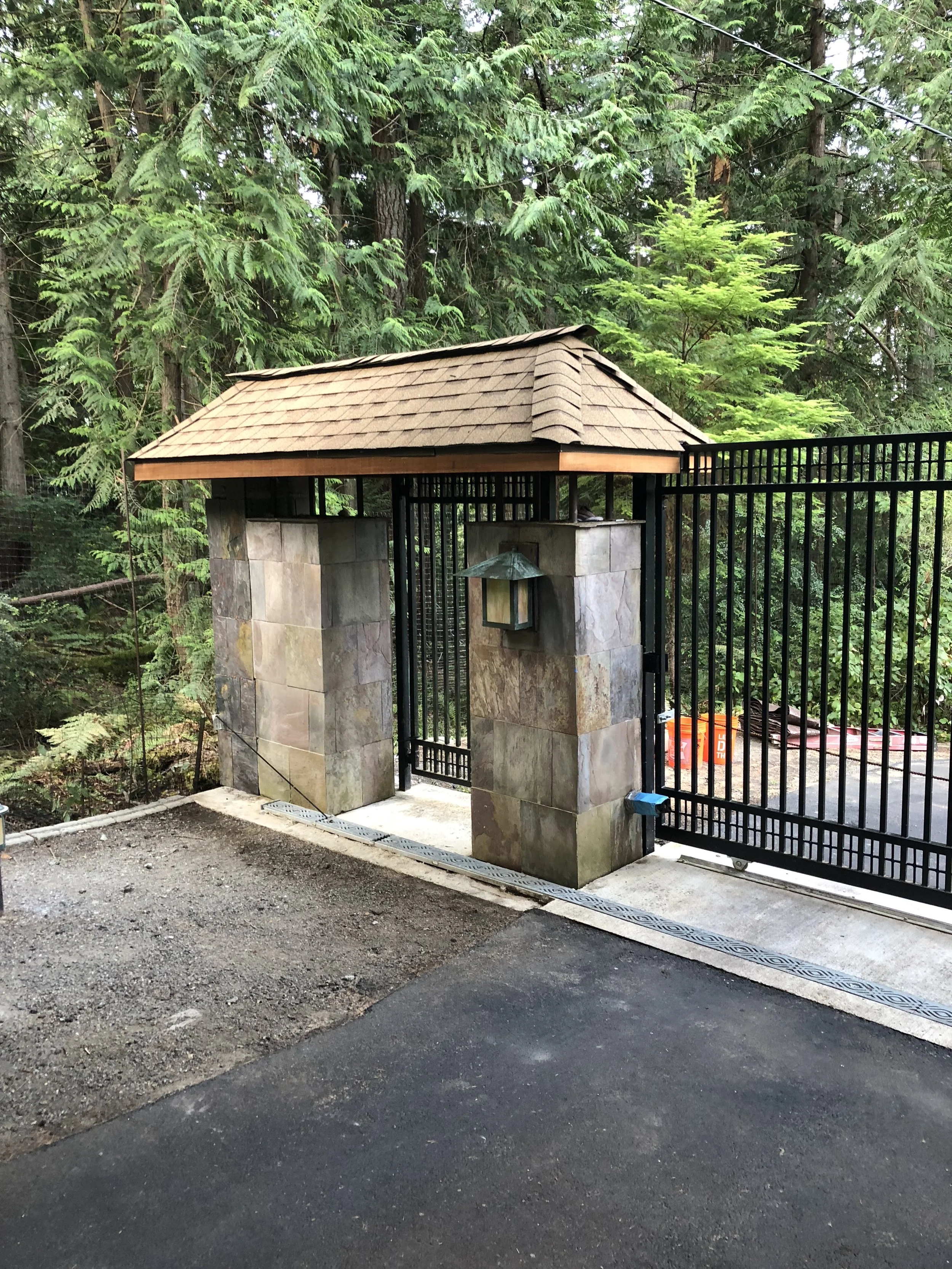 A stone pillar gate with a small roof and a lantern, surrounded by a black metal fence, with trees and dense foliage in the background.