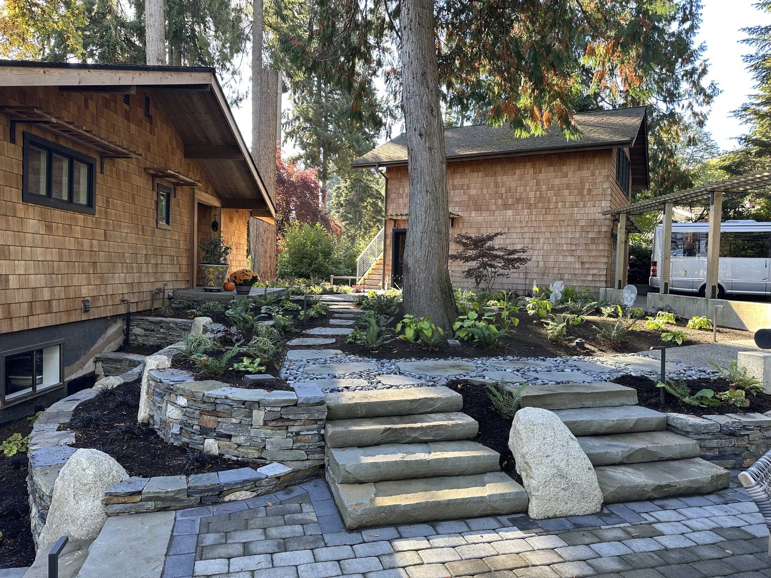 Outdoor scene with wooden houses, trees, and landscaped garden with pathway, stone steps, and plants.
