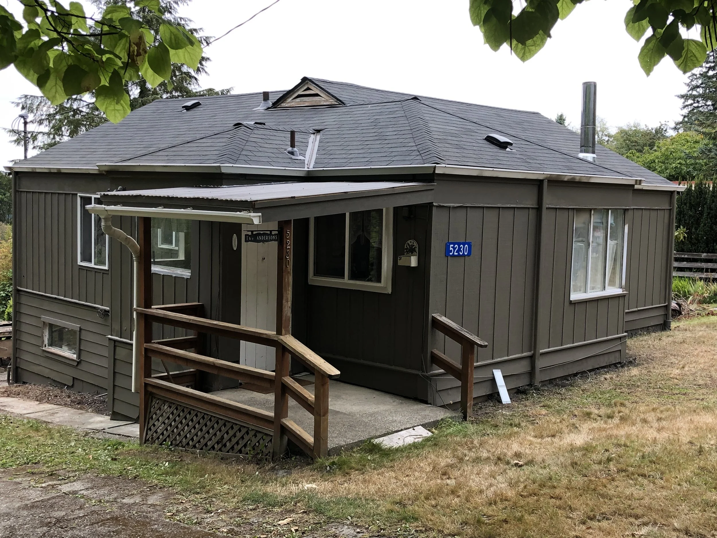 A house with gray exterior walls and a gray shingle roof surrounded by trees and greenery, with a deck and stairs leading to the entrance.
