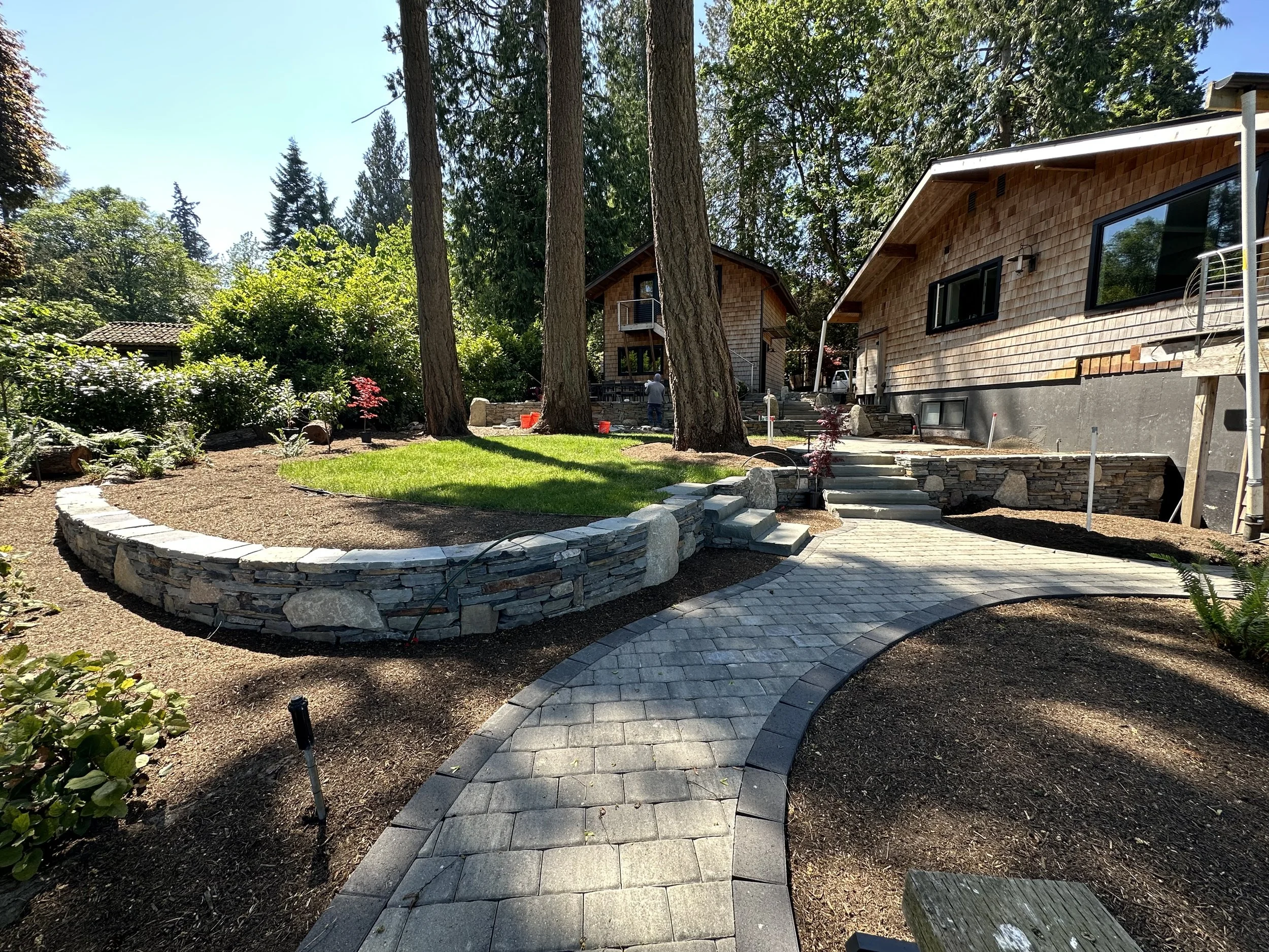 Backyard with trees, shrubs, a stone retaining wall, and a house with wooden shingles and large windows.