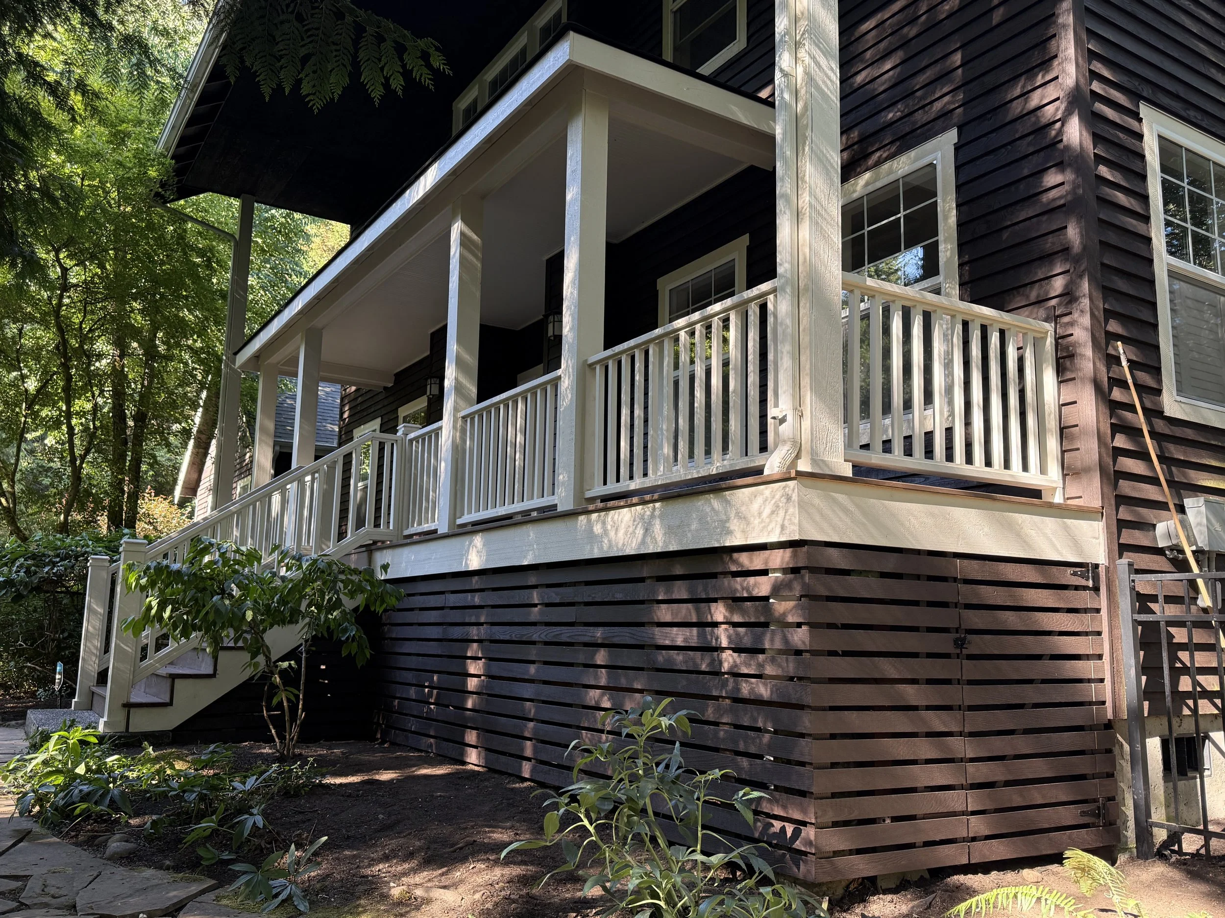 A house with dark brown wooden siding, white trim, and a balcony with white railing surrounded by green trees.