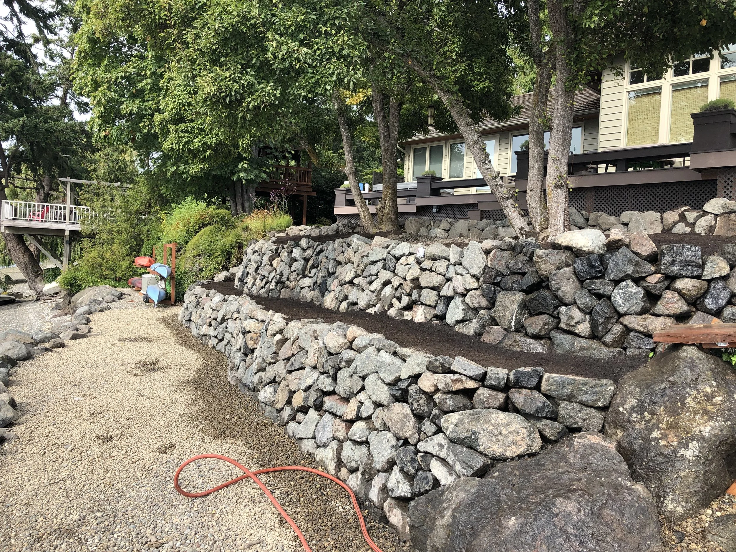 A backyard with a multi-level stone retaining wall, trees, and a deck attached to a house with large windows. Gardening tools and a hose are visible on the gravel ground.