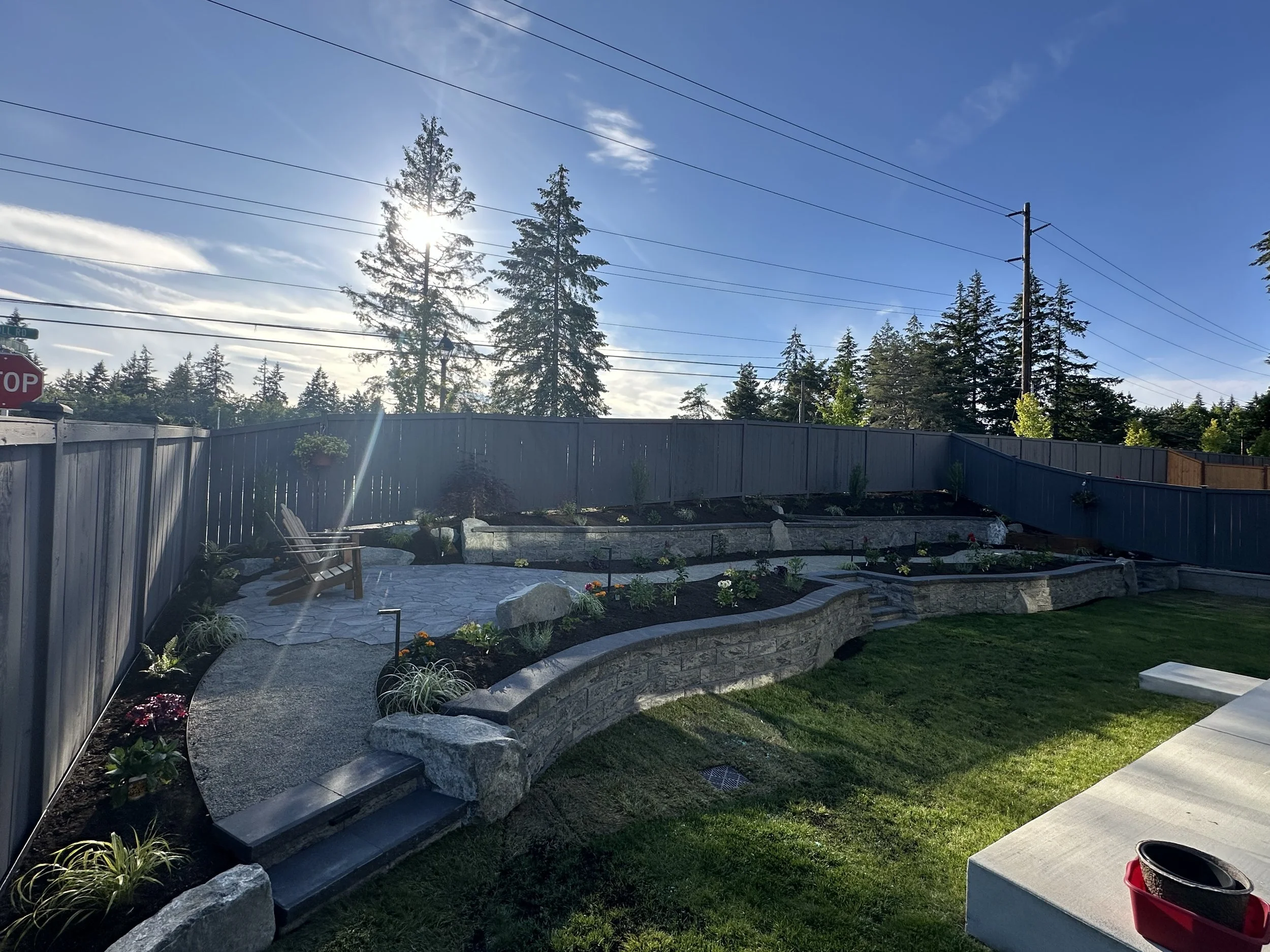 A backyard garden with a stone patio, garden beds, and a wooden fence. There are trees and power lines in the background, and the sky is partly cloudy with the sun shining through.