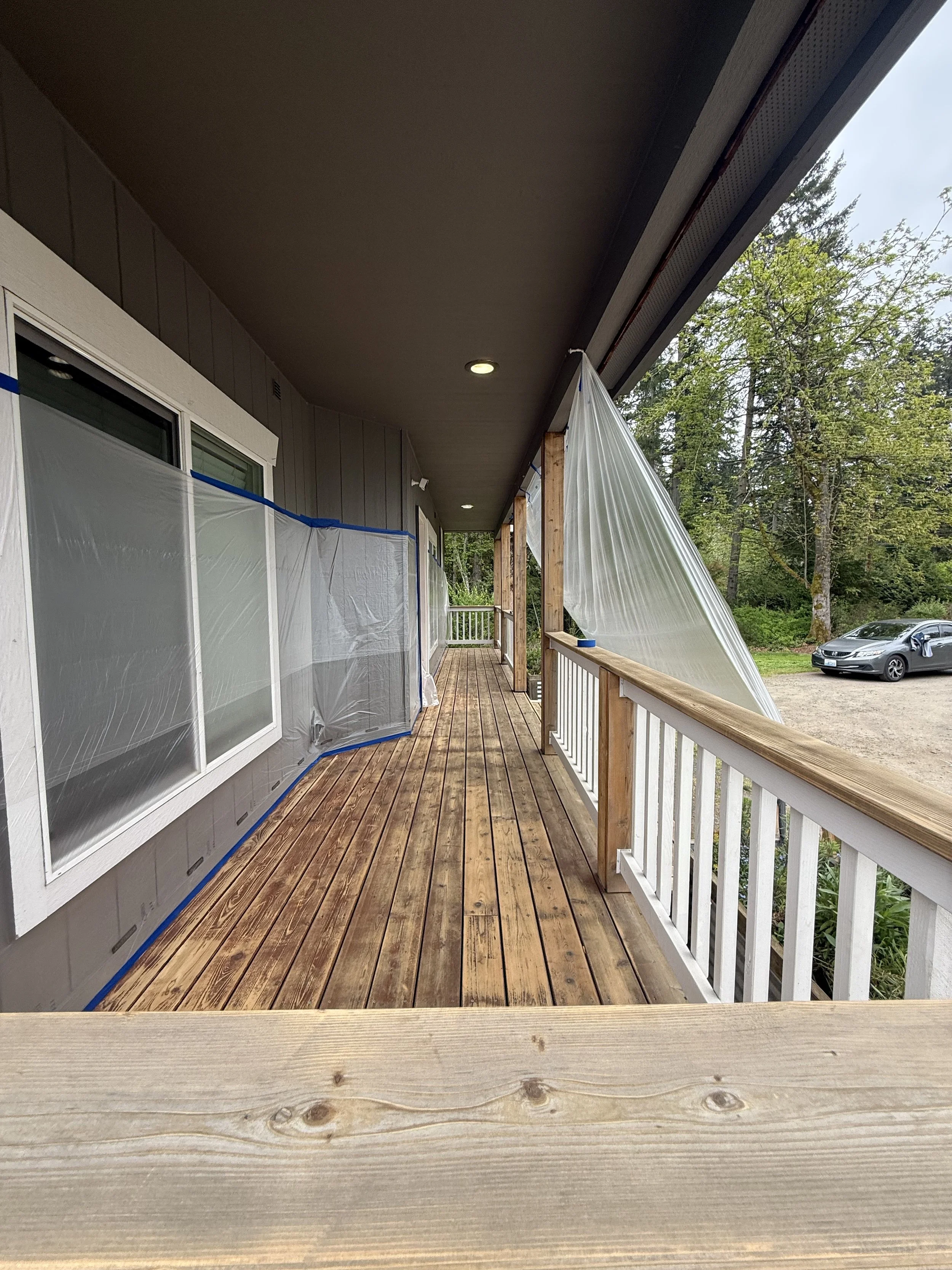 View of a balcony with wooden flooring, white railings, and a partially covered roof, with trees and cars visible in the background.