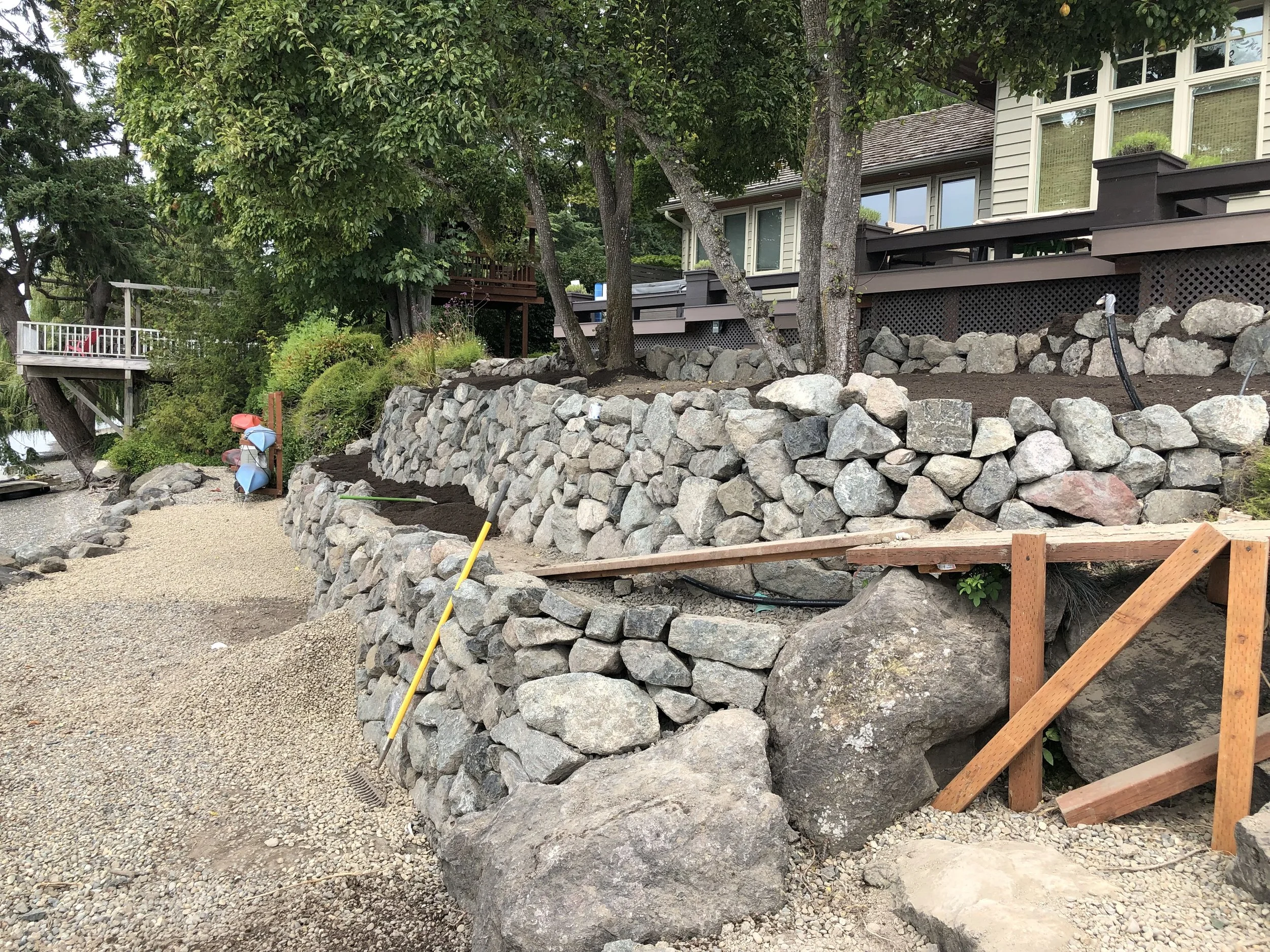 Residential backyard with a tiered landscape featuring large rocks, trees, and a house with a deck. There are construction tools and materials, including a yellow level and a rake, indicating ongoing landscaping work.