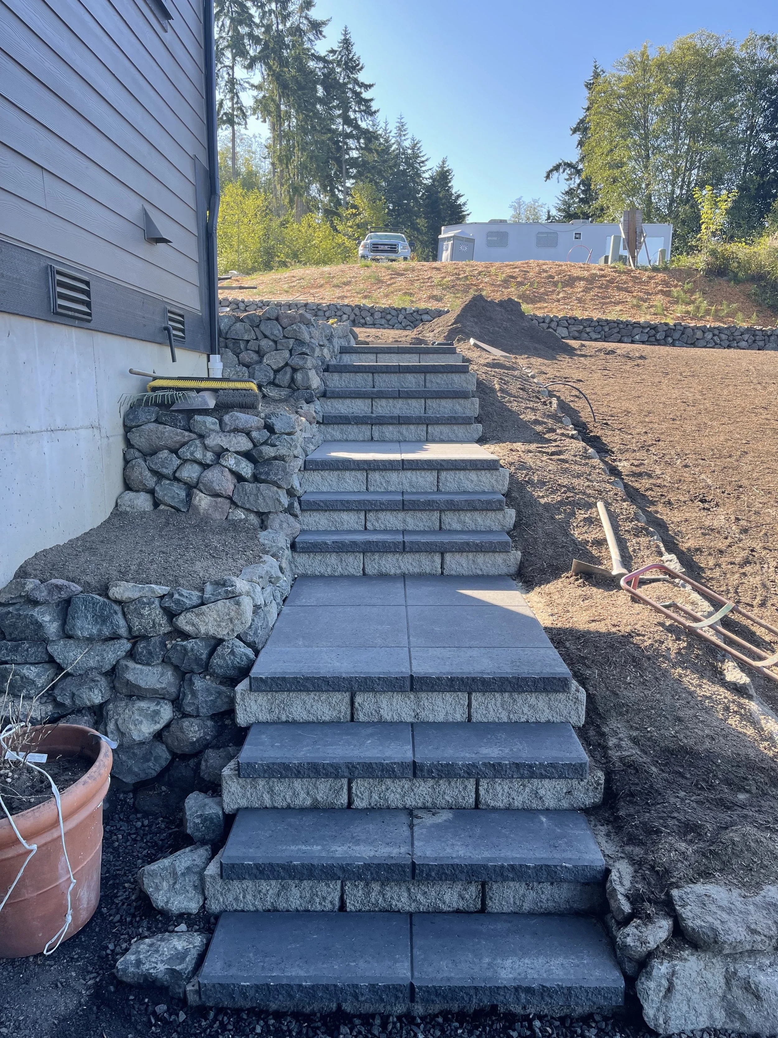Outdoor construction scene showing a set of stone steps leading up a slope, with rocks and dirt on either side, a large pot on the left, and a wheelbarrow on the right. In the background, there is a white vehicle and a white construction trailer unde