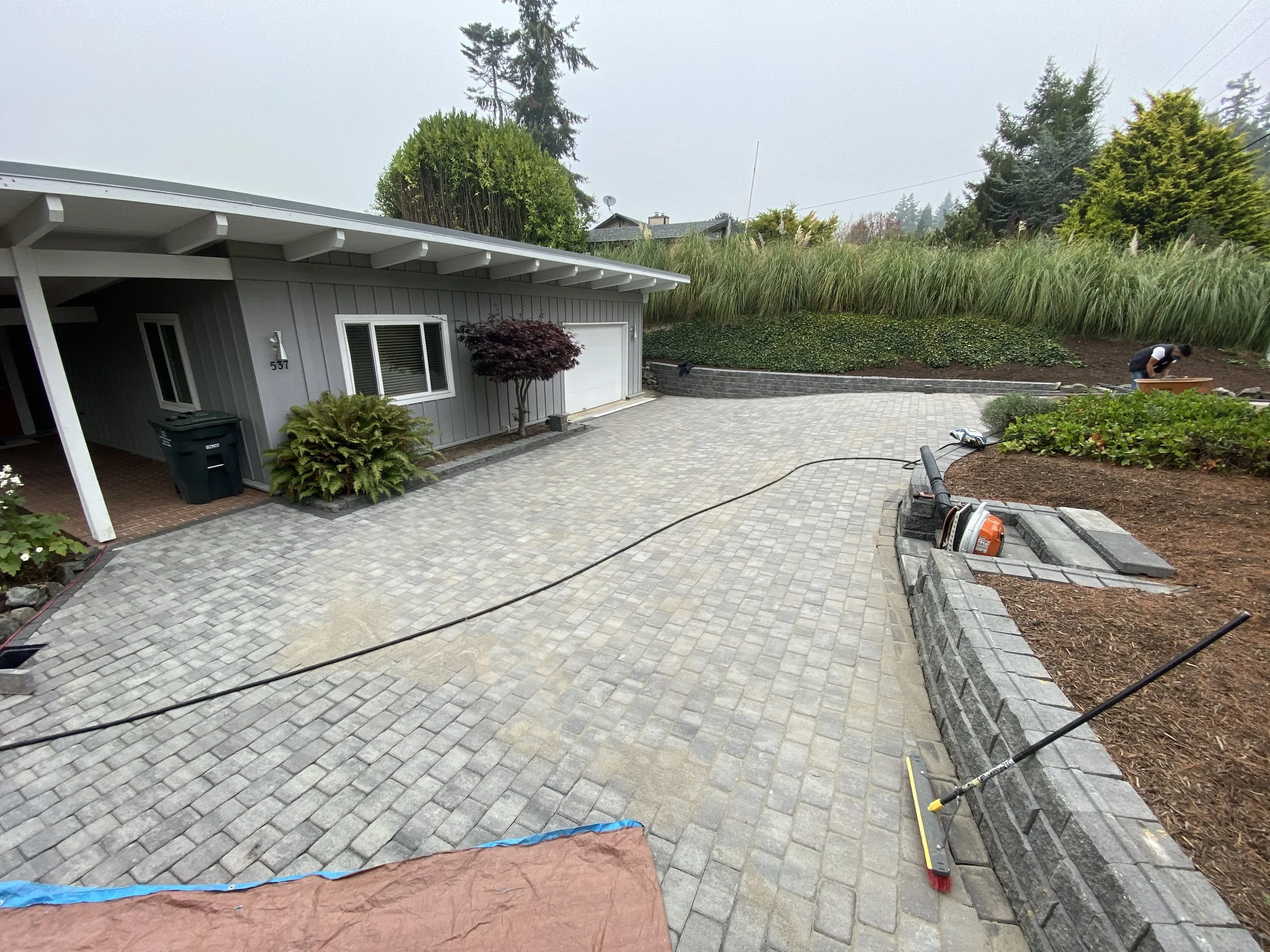A residential backyard showing a paved stone patio, landscaping, a garden worker, and house in the background with some fog or mist in the sky.