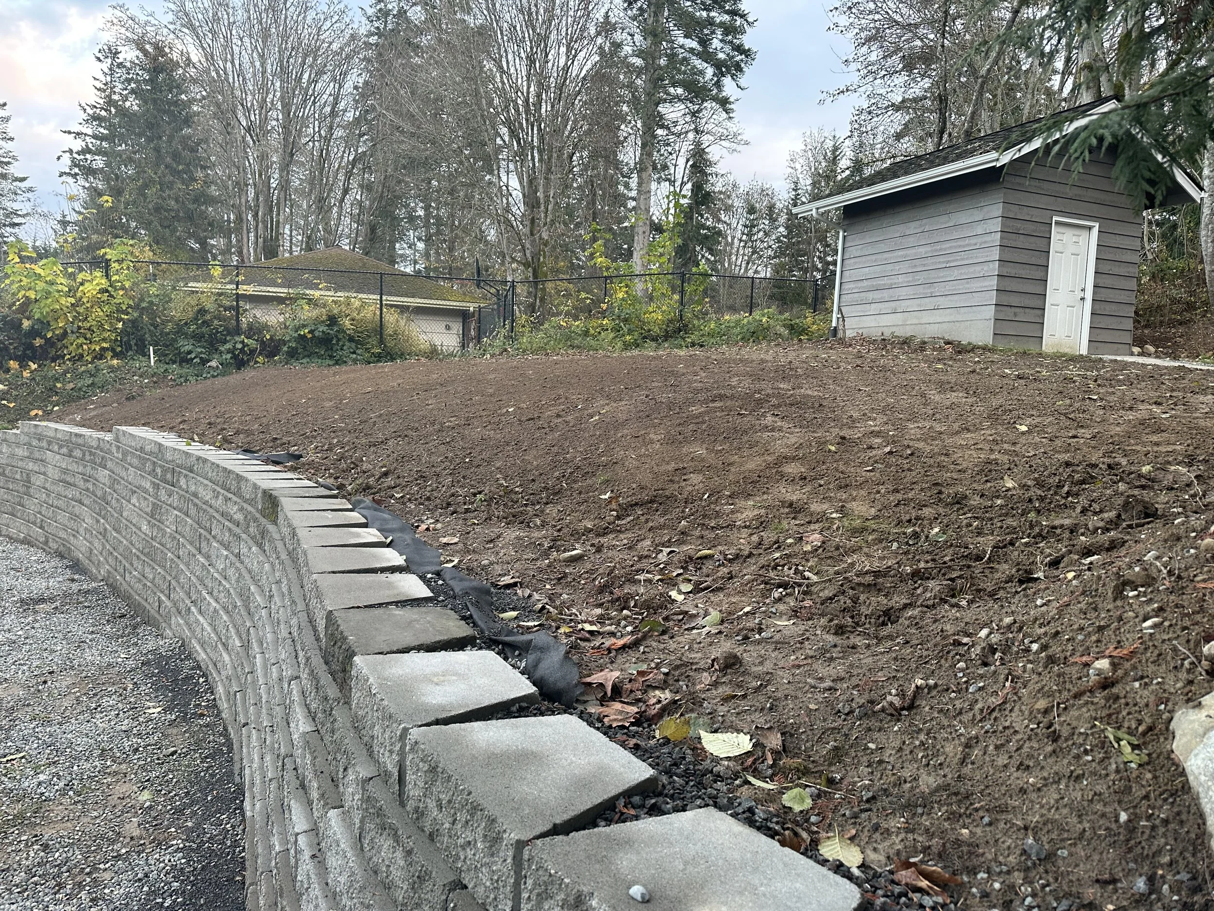 A backyard garden area with a freshly built stone retaining wall, empty soil bed, and a small gray shed in the background, surrounded by tall trees with sparse leaves.