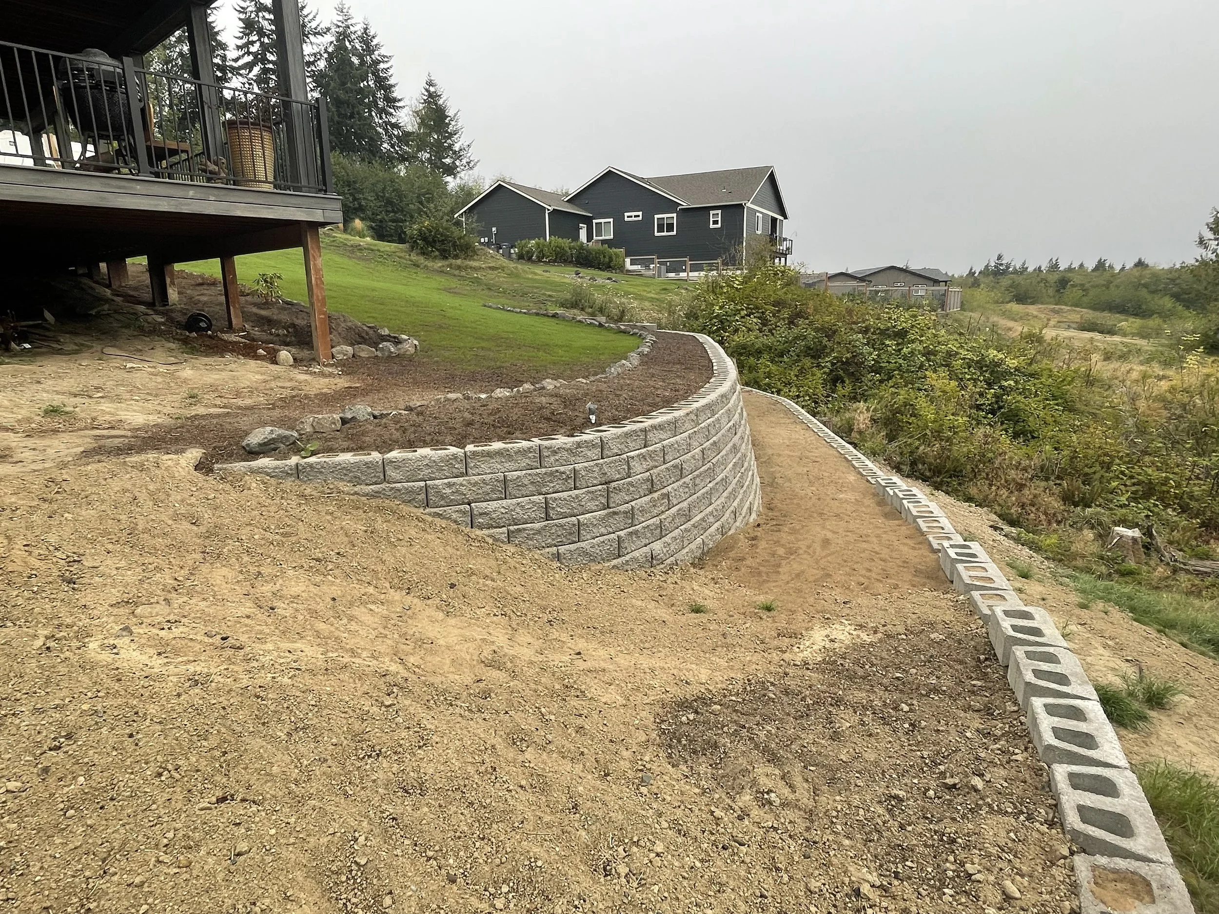 Construction site with a curved retaining wall made of gray concrete blocks, dirt pathway alongside, new houses in background, on a cloudy day.