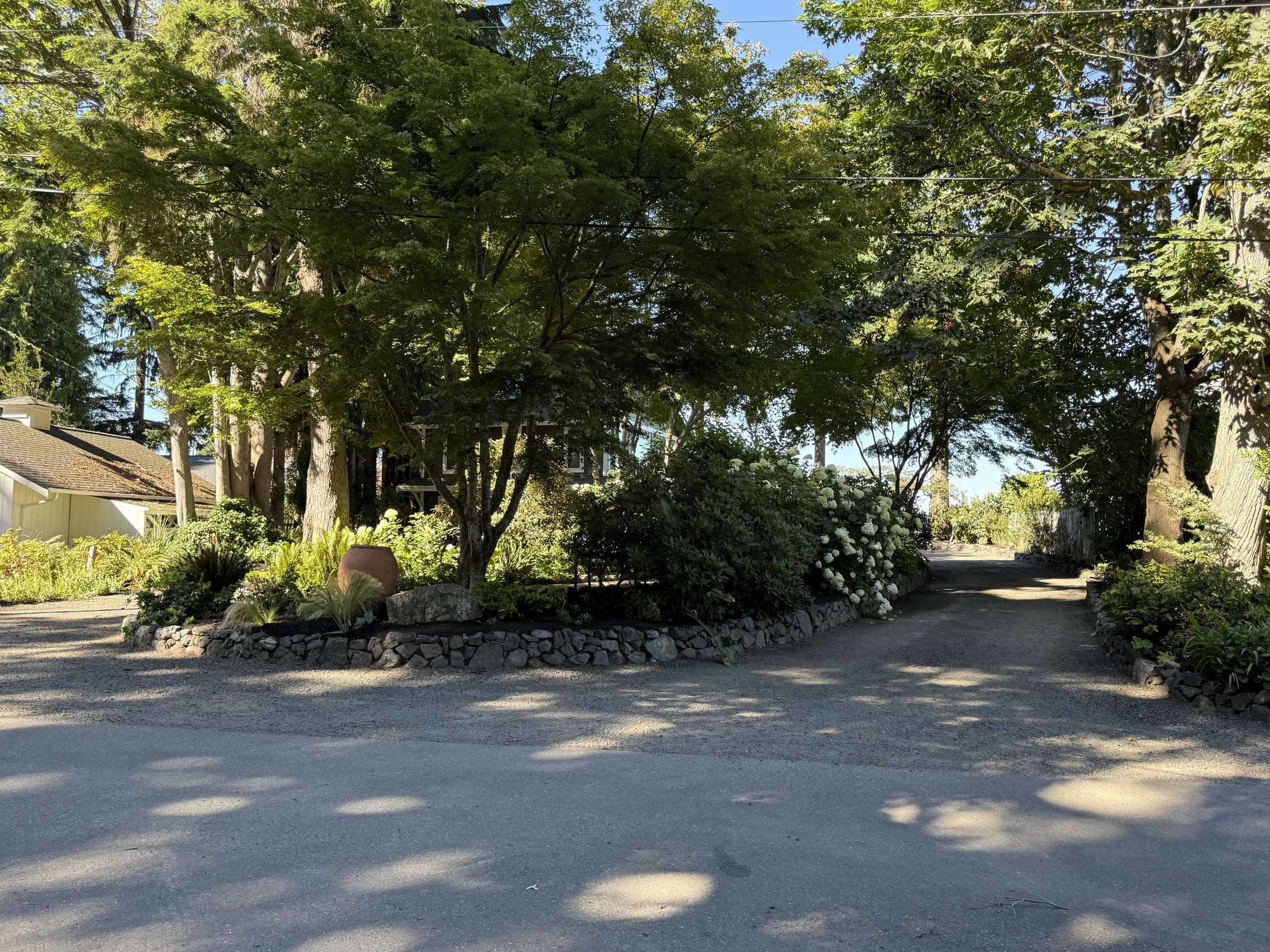 A garden scene with a gravel pathway, lush green trees, flowering shrubs, and a stone border around the planted area, with a house partially visible on the left and overhead power lines.