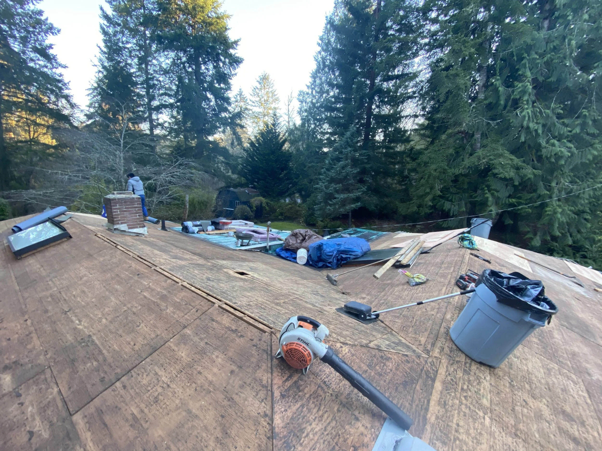 The image shows the roof of a house in the process of being re-shingled, with various roofing tools, materials, and equipment scattered across the roof surface. There are trees in the background.