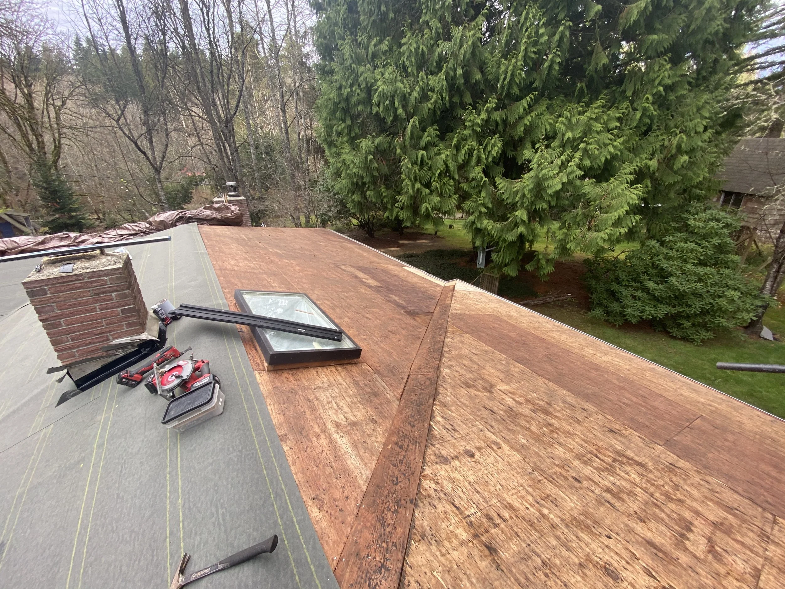The image shows the view from a rooftop with tools and equipment, including a cordless drill, a handsaw, and a toolbox, positioned near a chimney. The roof is partially covered with a tarp, and the background features tall, leafless trees and a large
