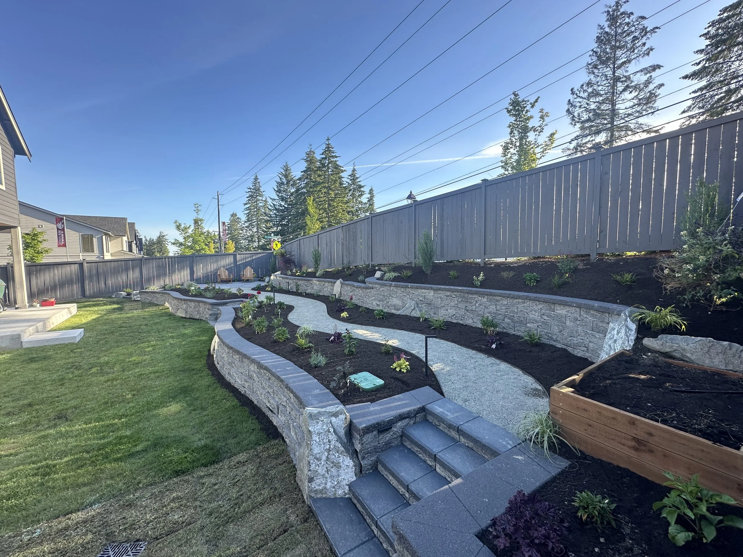 A landscaped backyard with a stone-edged garden bed filled with various plants, a winding concrete pathway, and a green lawn. A gray wooden fence runs along the back, with tall evergreen trees behind it. Houses with beige siding are visible on the le