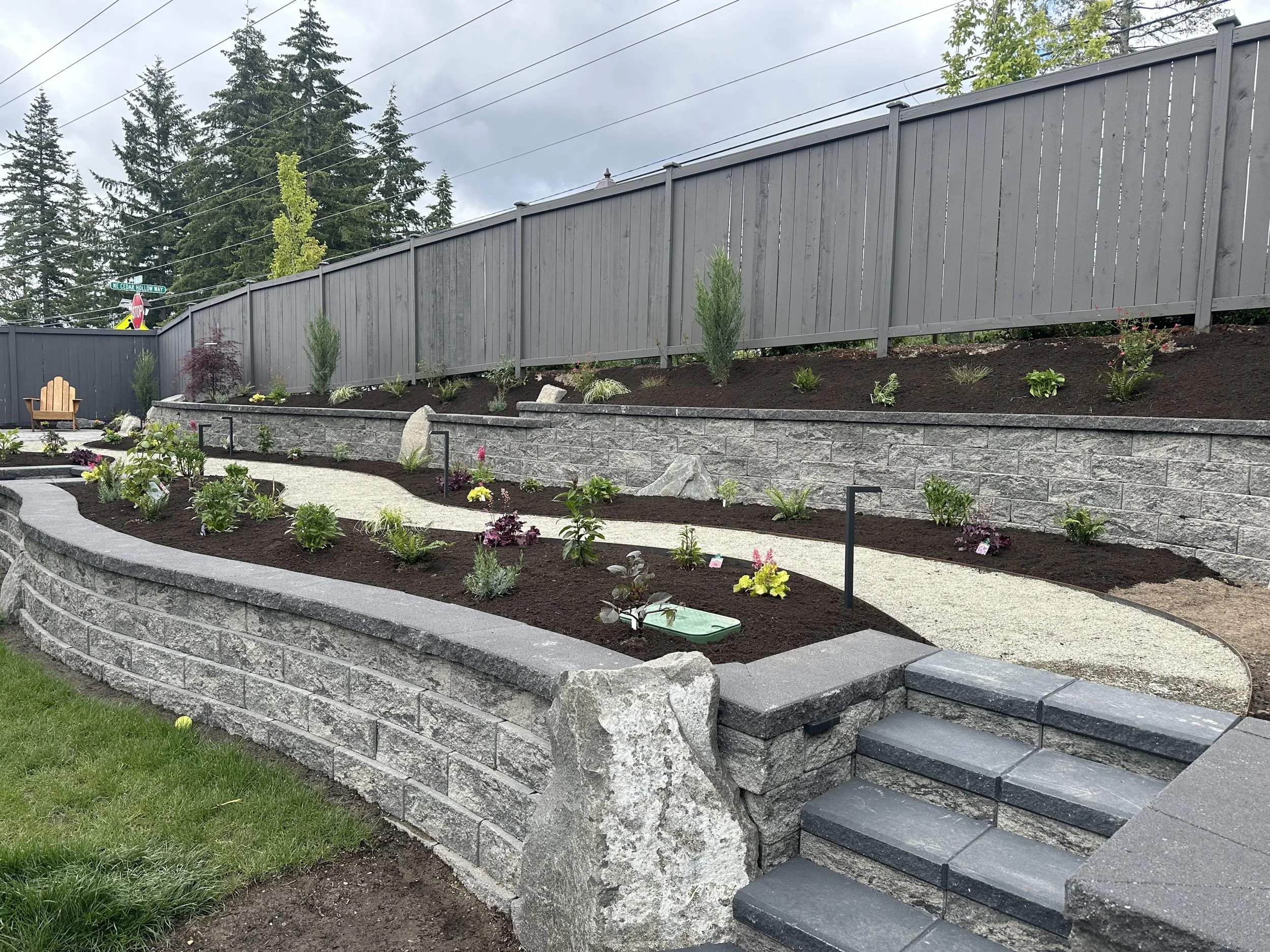A landscaped backyard with a two-tiered stone retaining wall, planting beds with various small plants and bushes, a curved concrete pathway, and a tall gray wooden fence. There are trees and power lines in the background under an overcast sky.